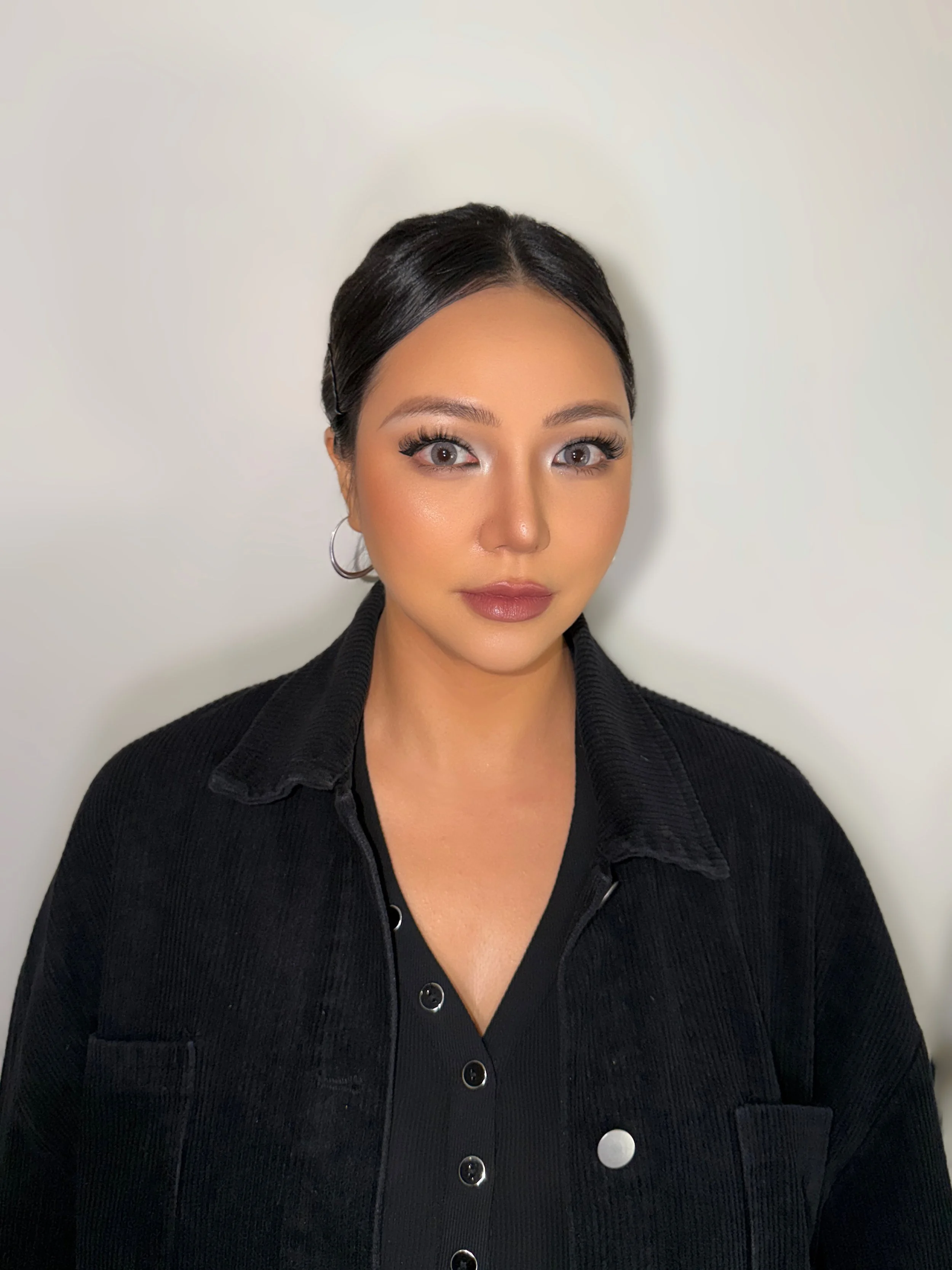 Close-up portrait of a young woman with dark hair, wearing hoop earrings and a black button-up top, standing against a plain light-colored background.