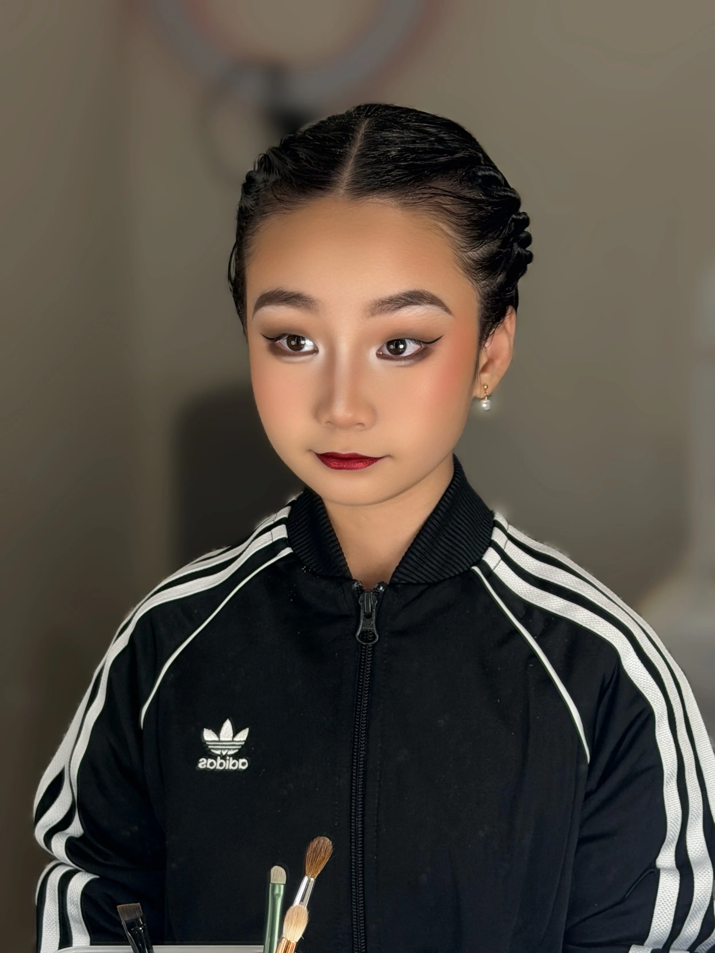 A young woman with dark, styled hair and makeup, wearing a black adidas tracksuit, holding makeup brushes.