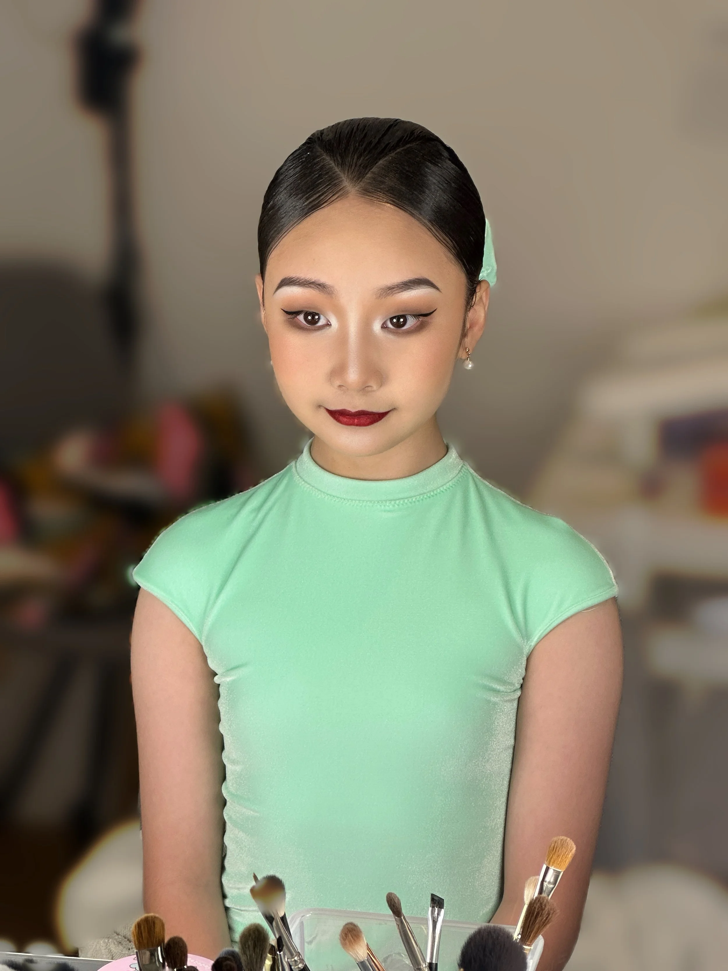 A young woman with styled hair, wearing a light green top, and makeup, sitting at a table with various makeup brushes in front of her, with a blurred background.