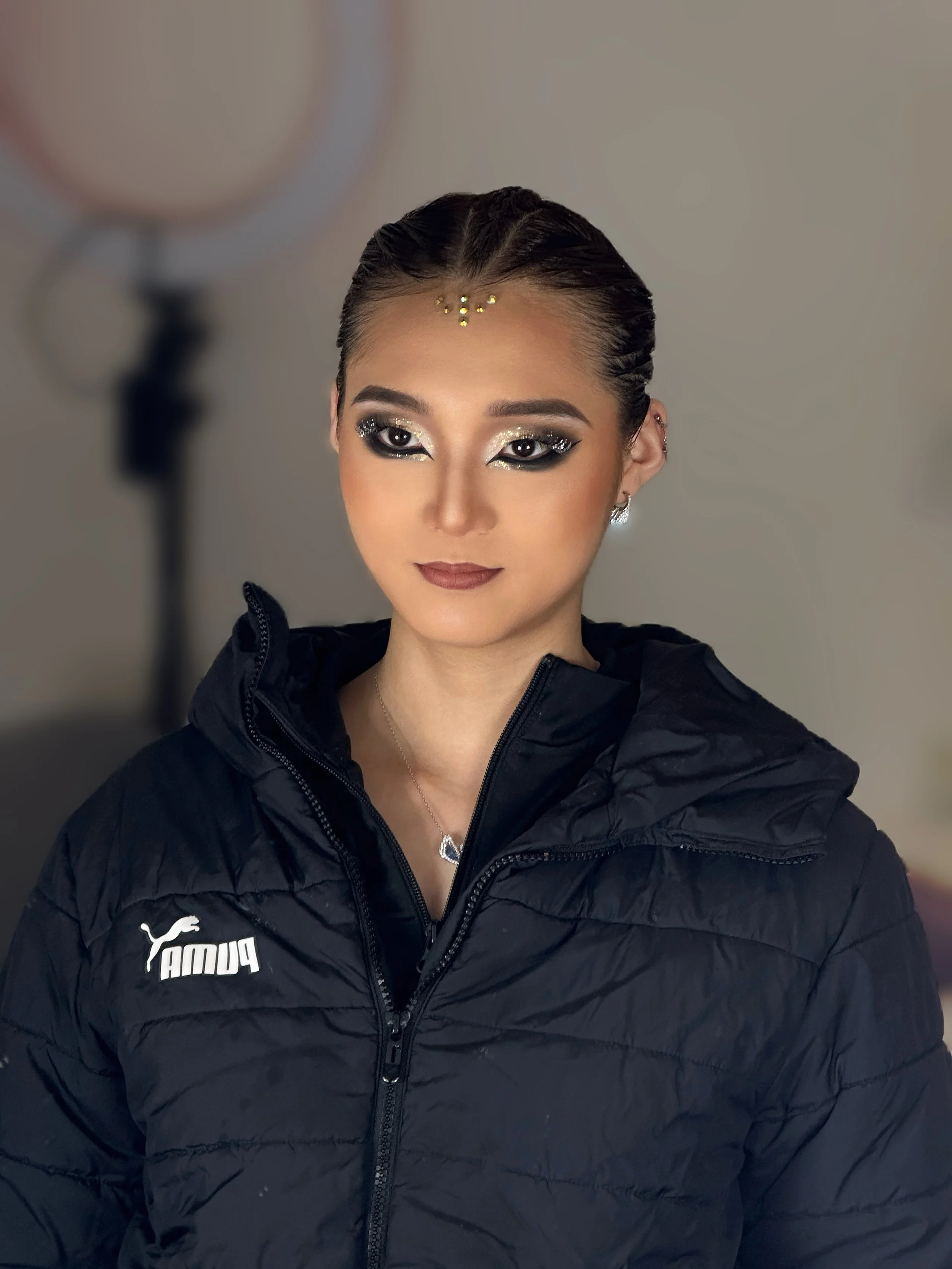 Young woman with braided hair and makeup featuring silver and black glitter eyeshadow, wearing a black Puma jacket and a necklace with a heart-shaped pendant, indoors with a blurred background.