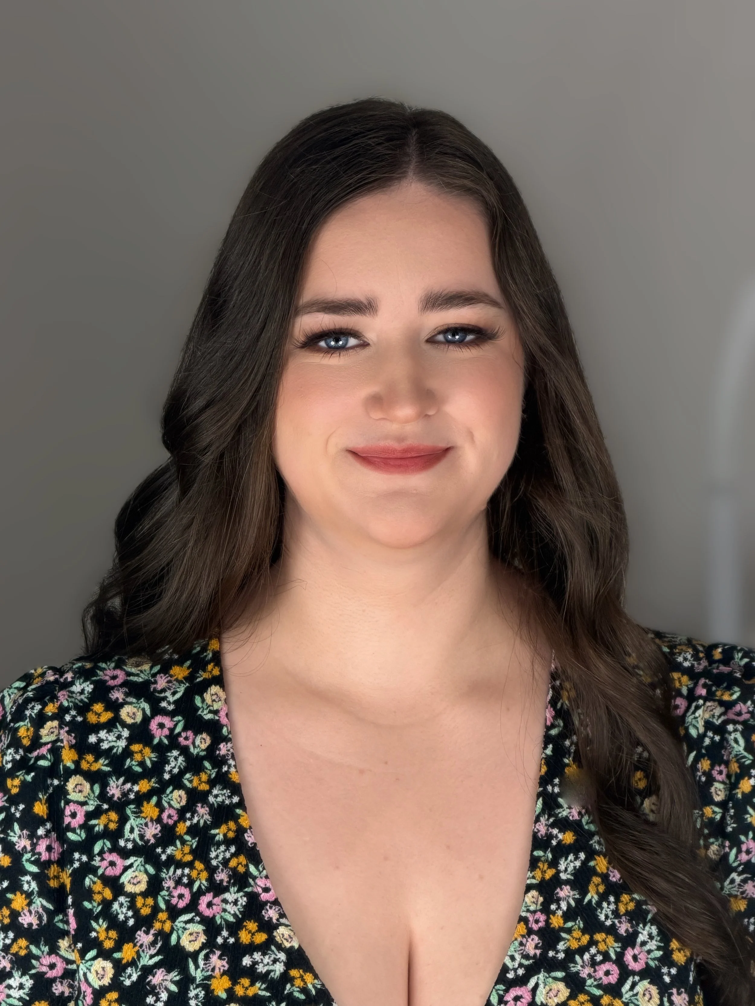 A woman with long brown hair, blue eyes, and makeup smiling, wearing a black floral dress with a V-neck. She is standing against a plain grey background.