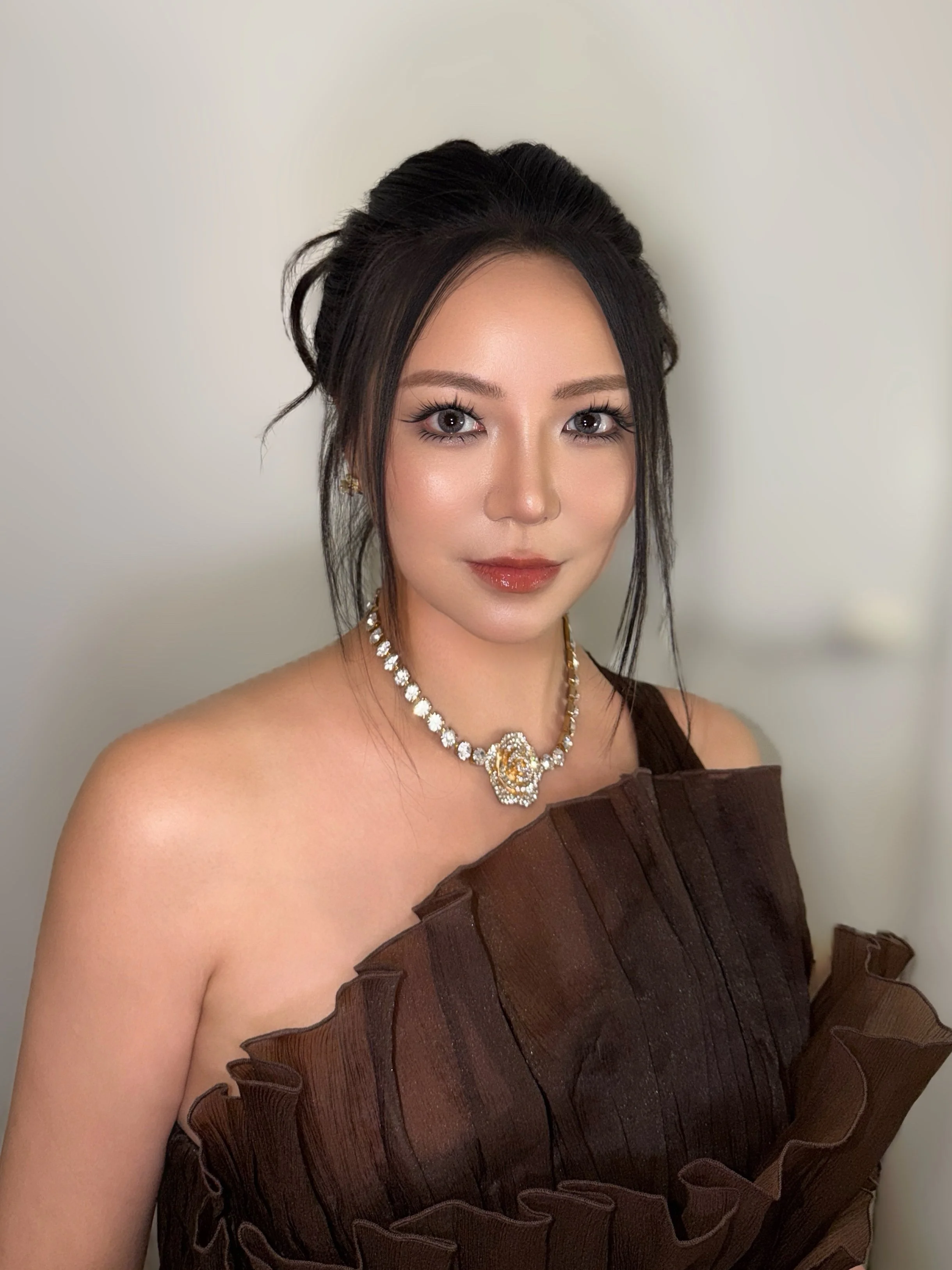 A woman with dark hair styled in an elegant updo, wearing a necklace with a large pendant and a brown dress with ruffled details, standing against a plain white wall.