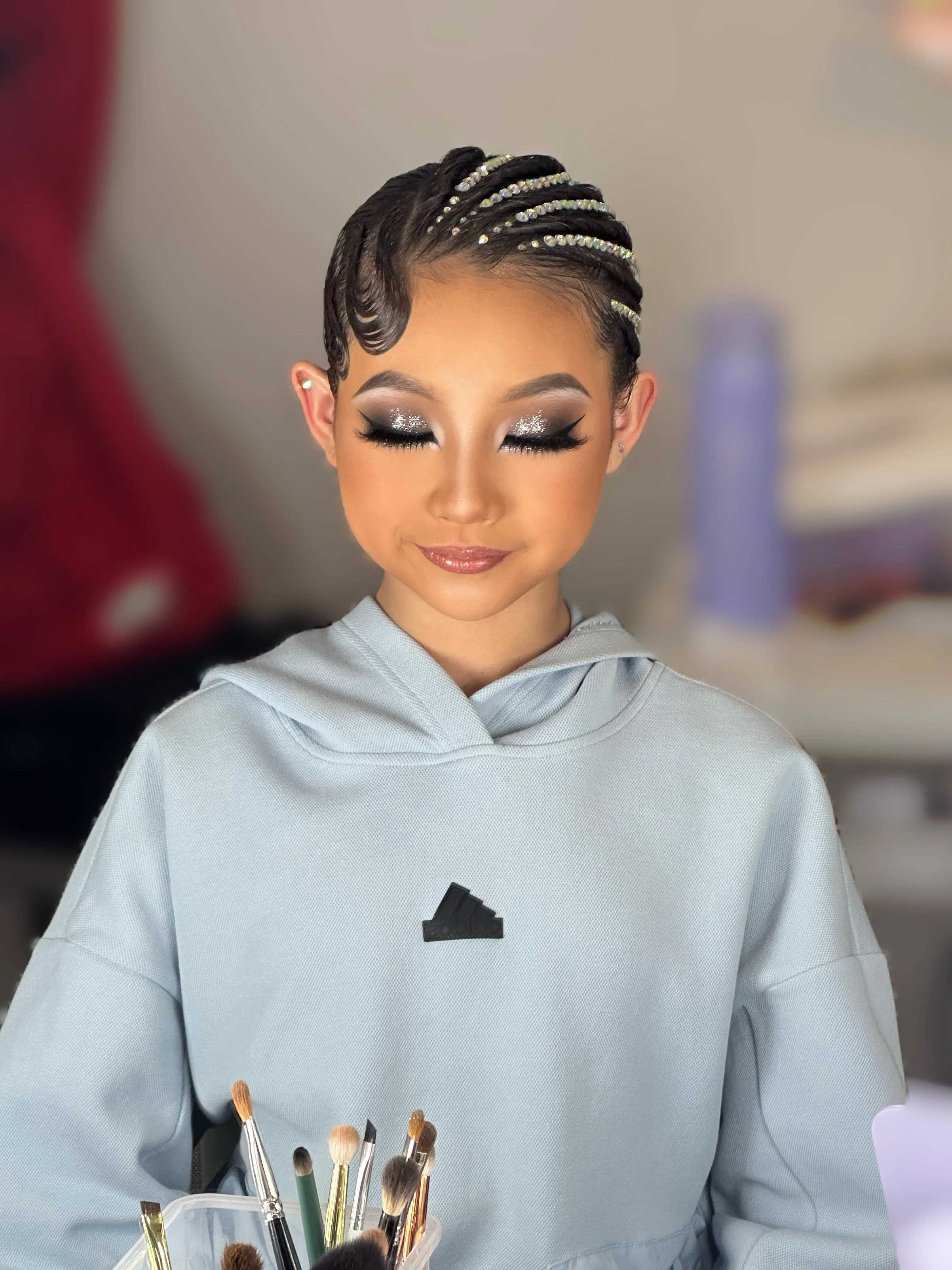 A young girl with elaborate hairstyle and makeup sitting in a room with makeup brushes in front of her.