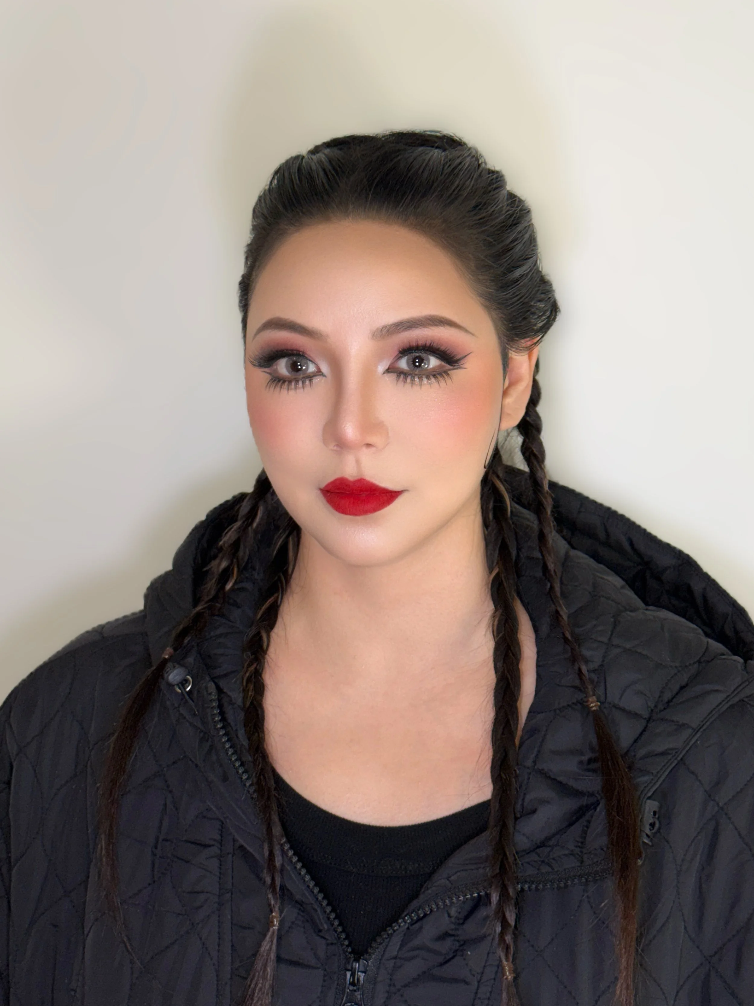 Close-up of a woman with braided hair, red lipstick, and eye makeup, wearing a black quilted jacket, against a plain light-colored background.