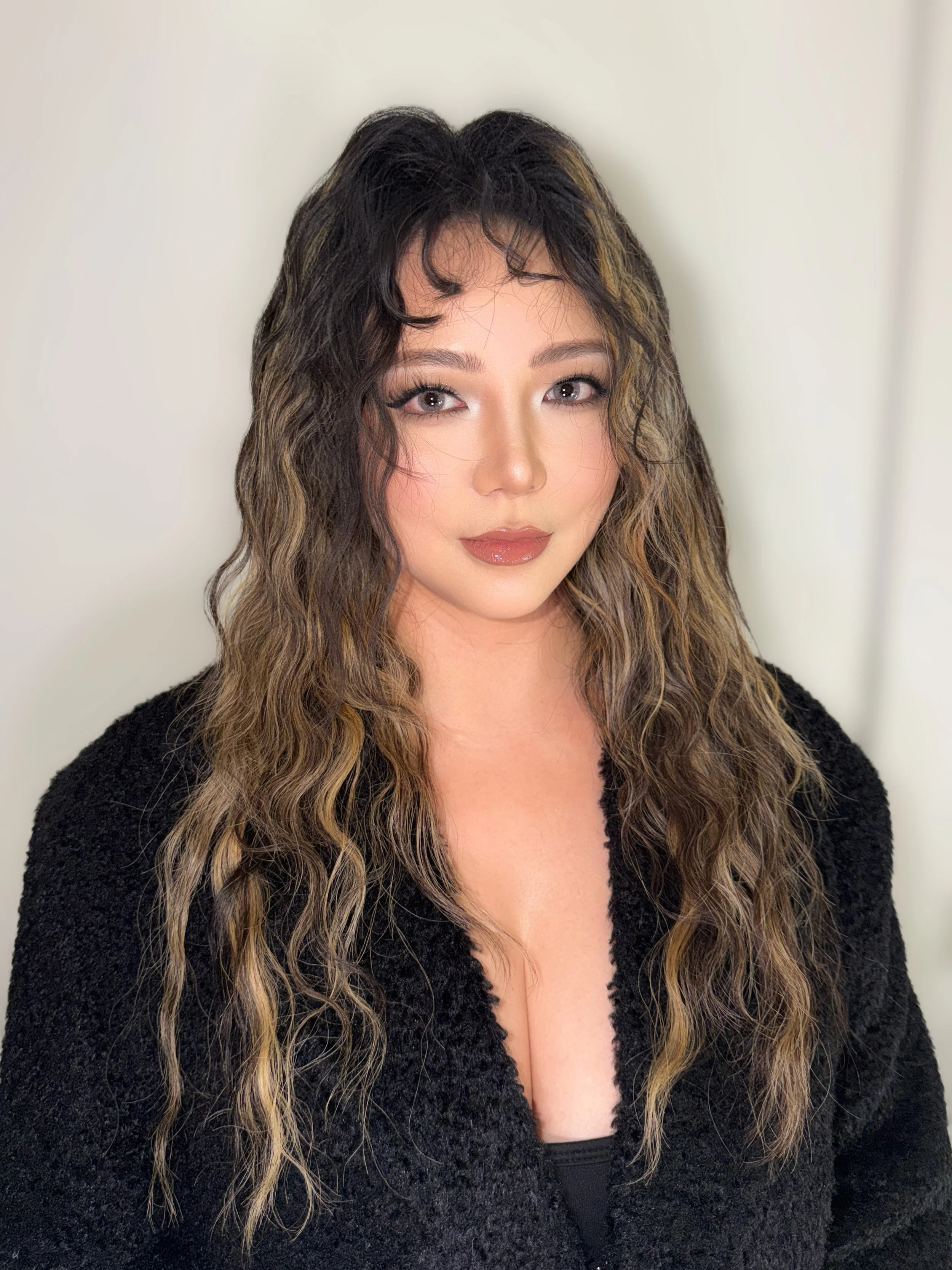 A woman with long, wavy brown hair and light makeup wearing a black top with a deep neckline, standing against a plain light-colored wall.