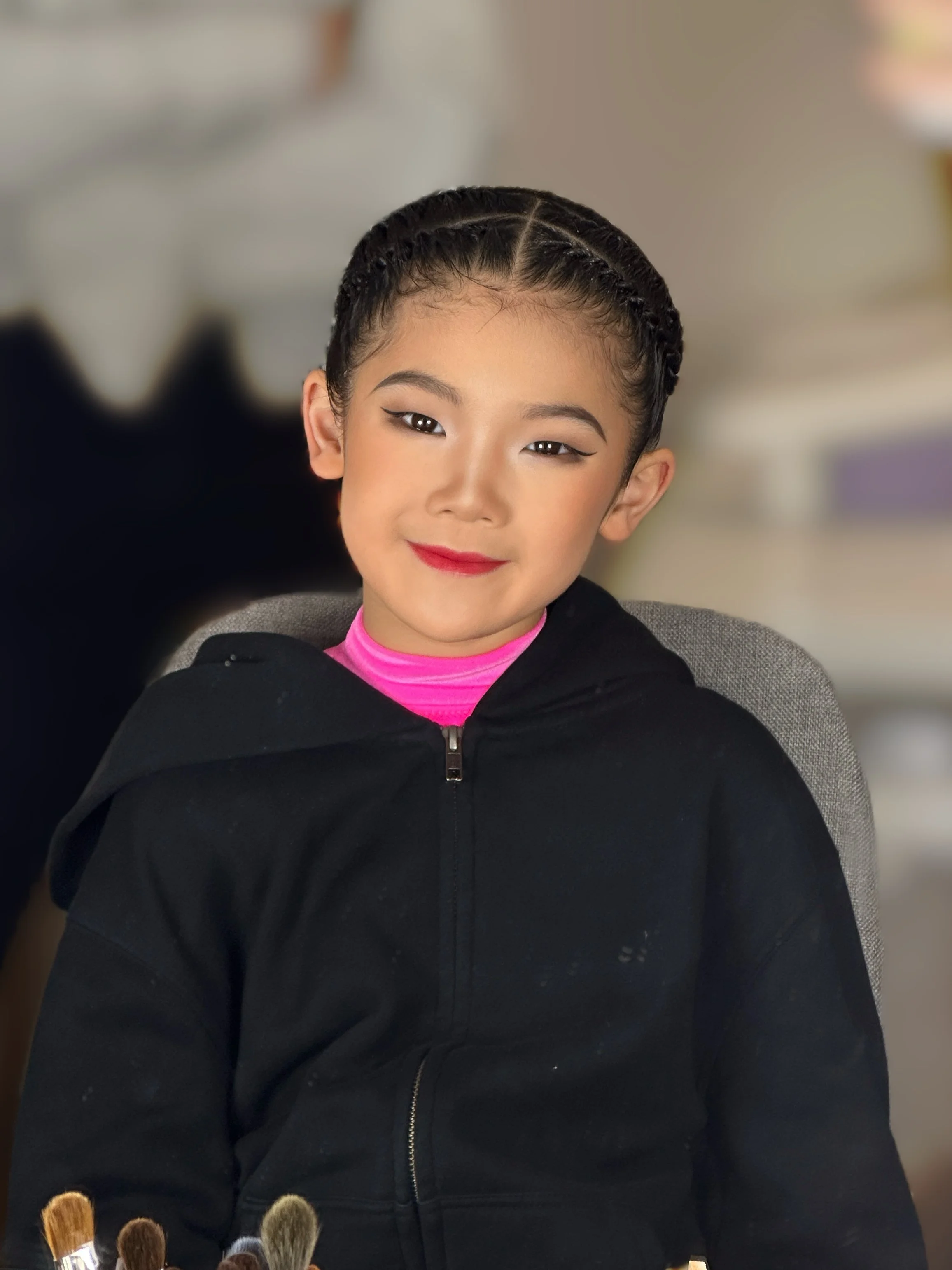 Young girl with braided hair, wearing a black jacket and pink shirt, sitting on a grey chair in an indoor setting.