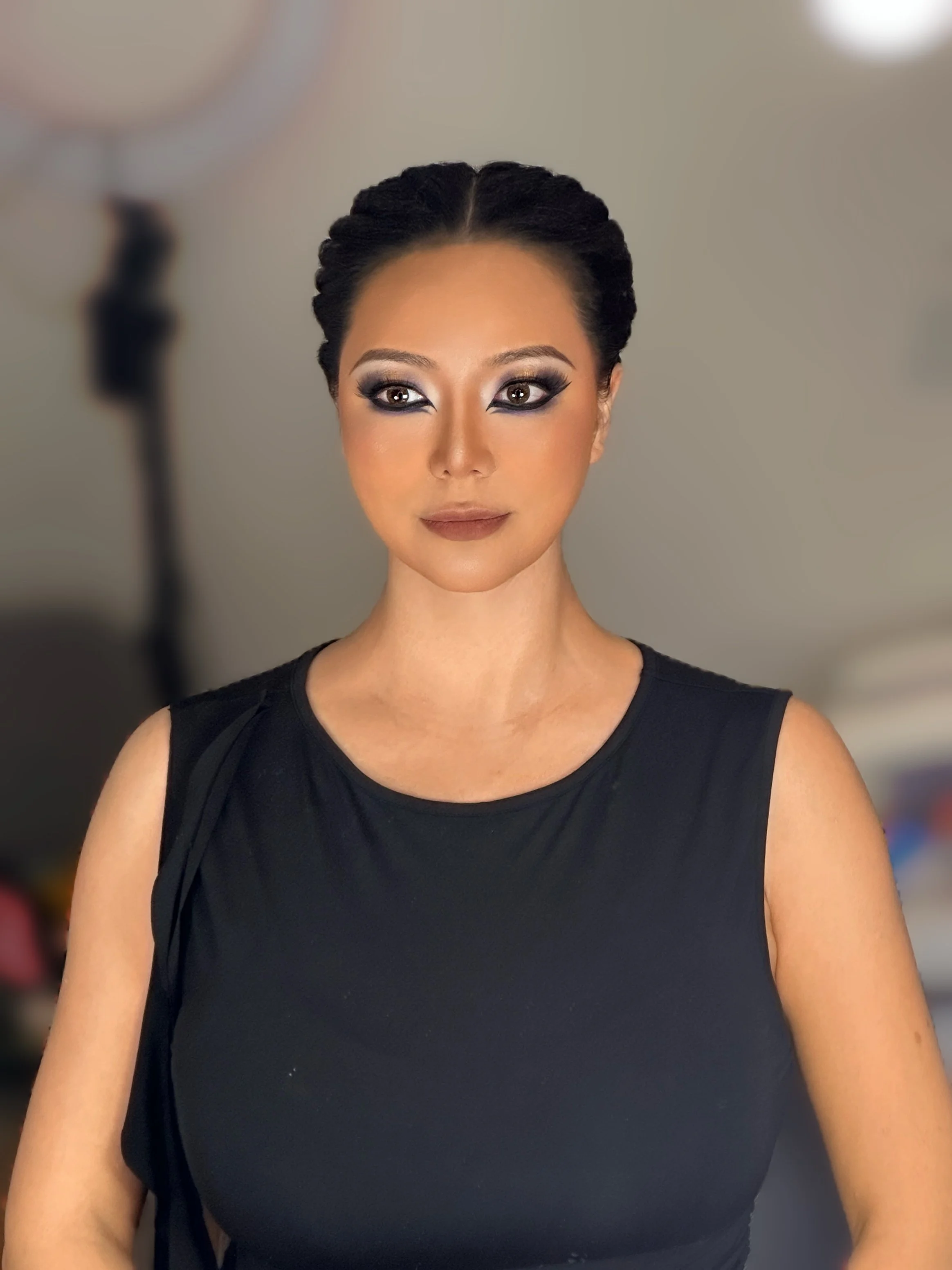 A woman with styled dark hair and dramatic makeup, wearing a sleeveless black top, posing indoors with studio lighting in the background.