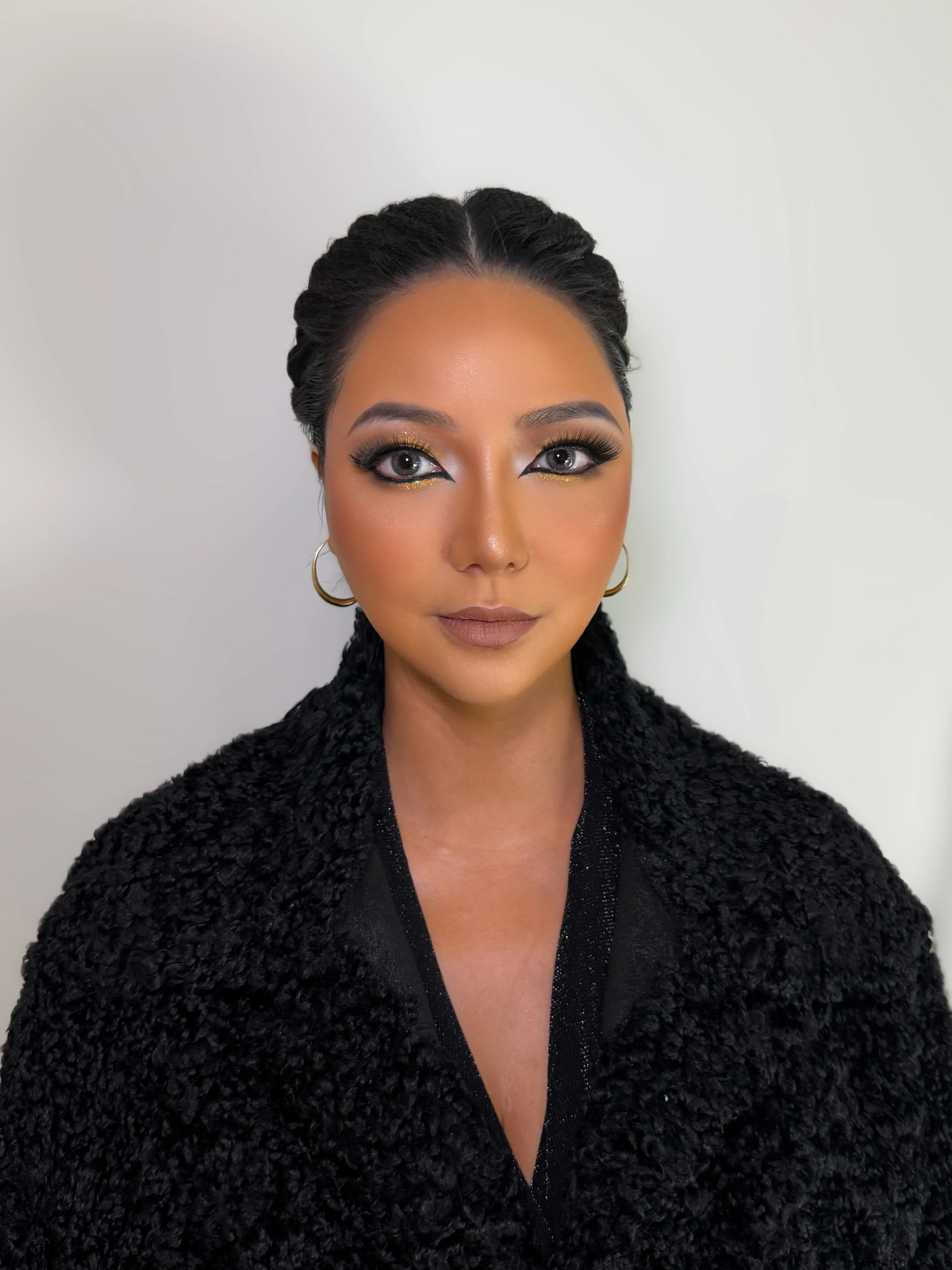 A woman with light purple eyeshadow, long thick eyelashes, and gold eyeliner beneath her eyes. She has dark hair styled in twists or cornrows, wears large gold hoop earrings, and a black fuzzy coat over a sparkly black top. The background is plain wh