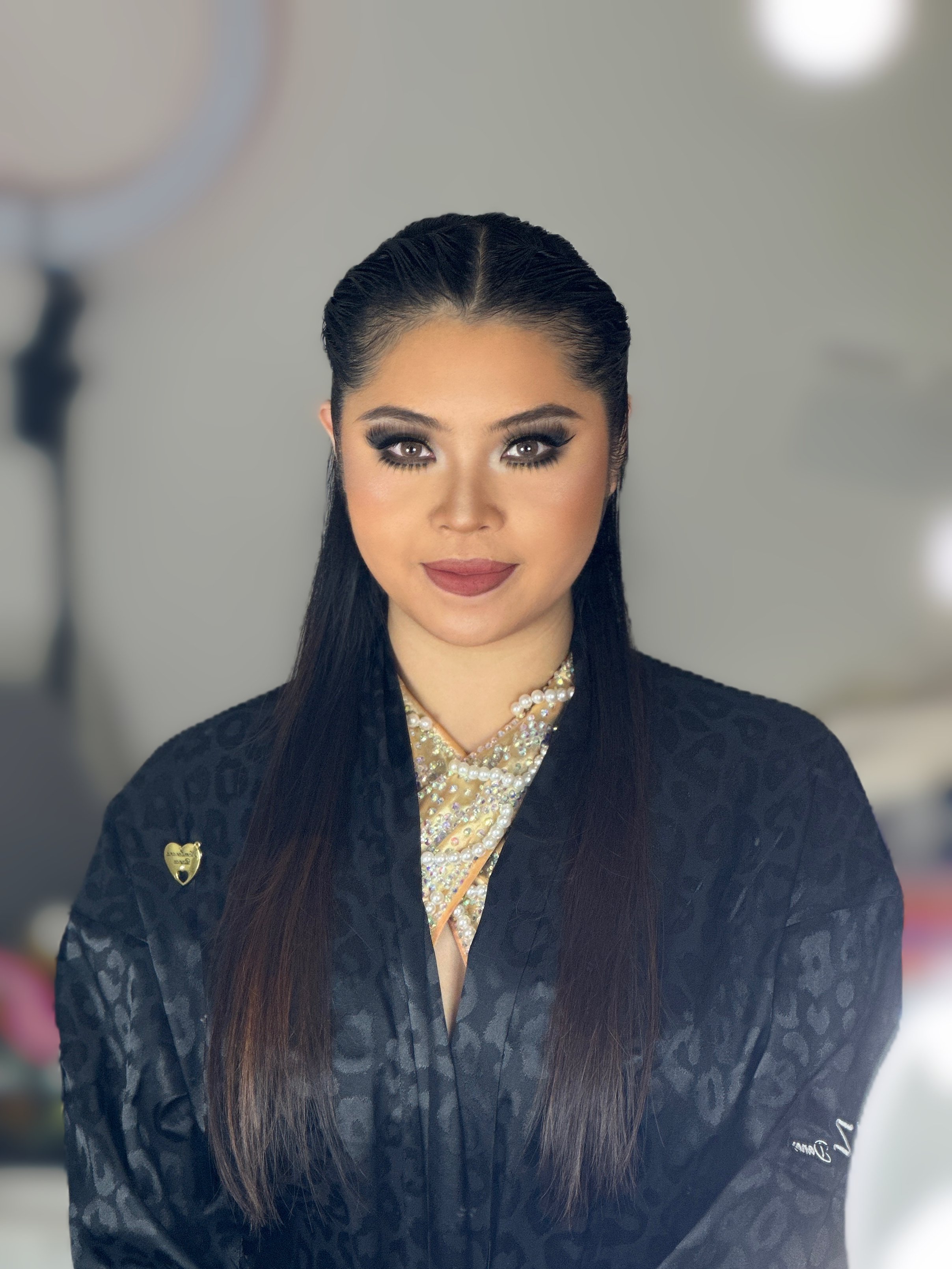 A young woman with dark hair, makeup, and jewelry, wearing a dark leopard print jacket, posing indoors.