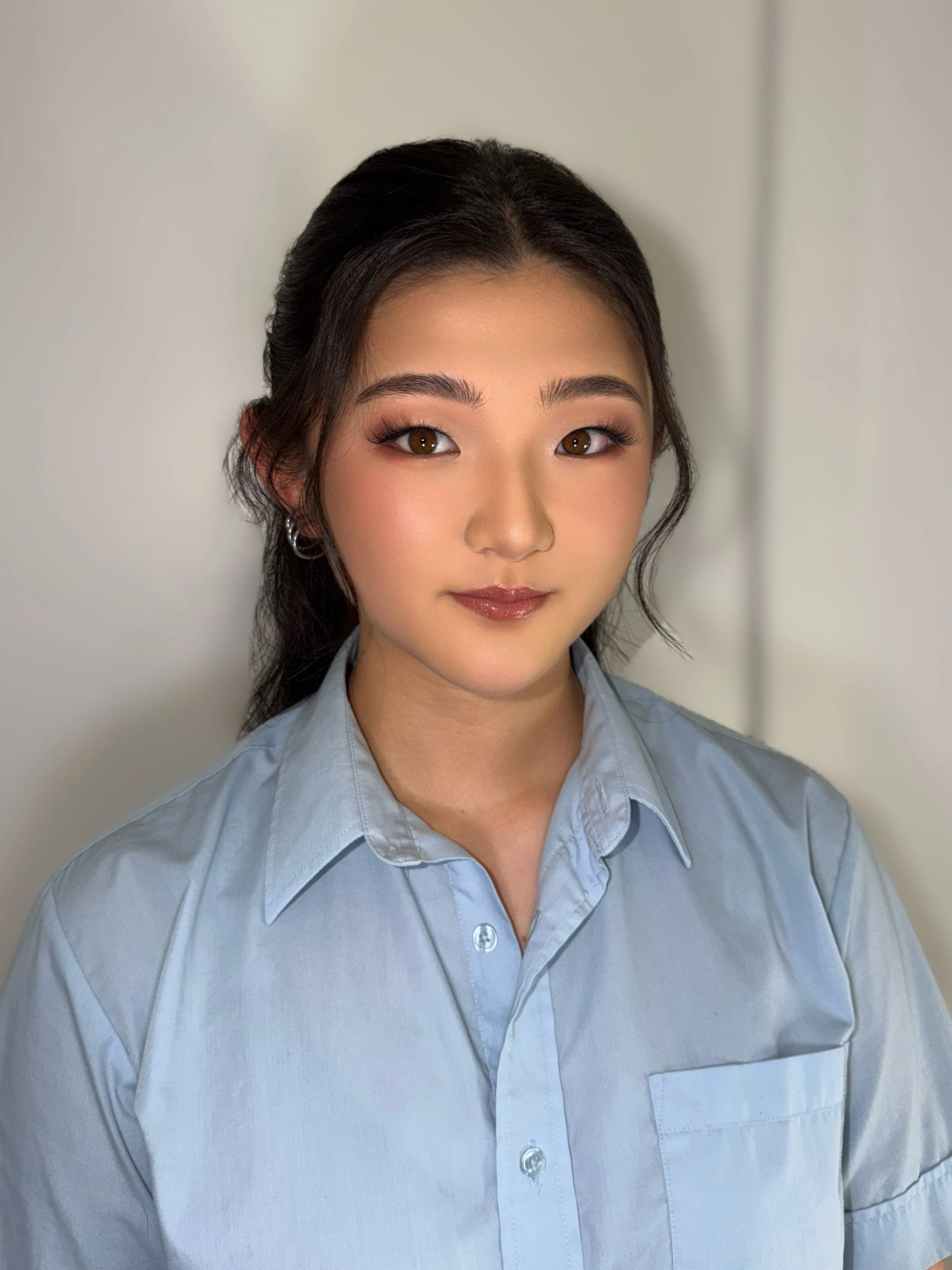 Close-up of a young woman with dark hair styled back, wearing a light blue collared shirt, standing in front of a plain neutral background.