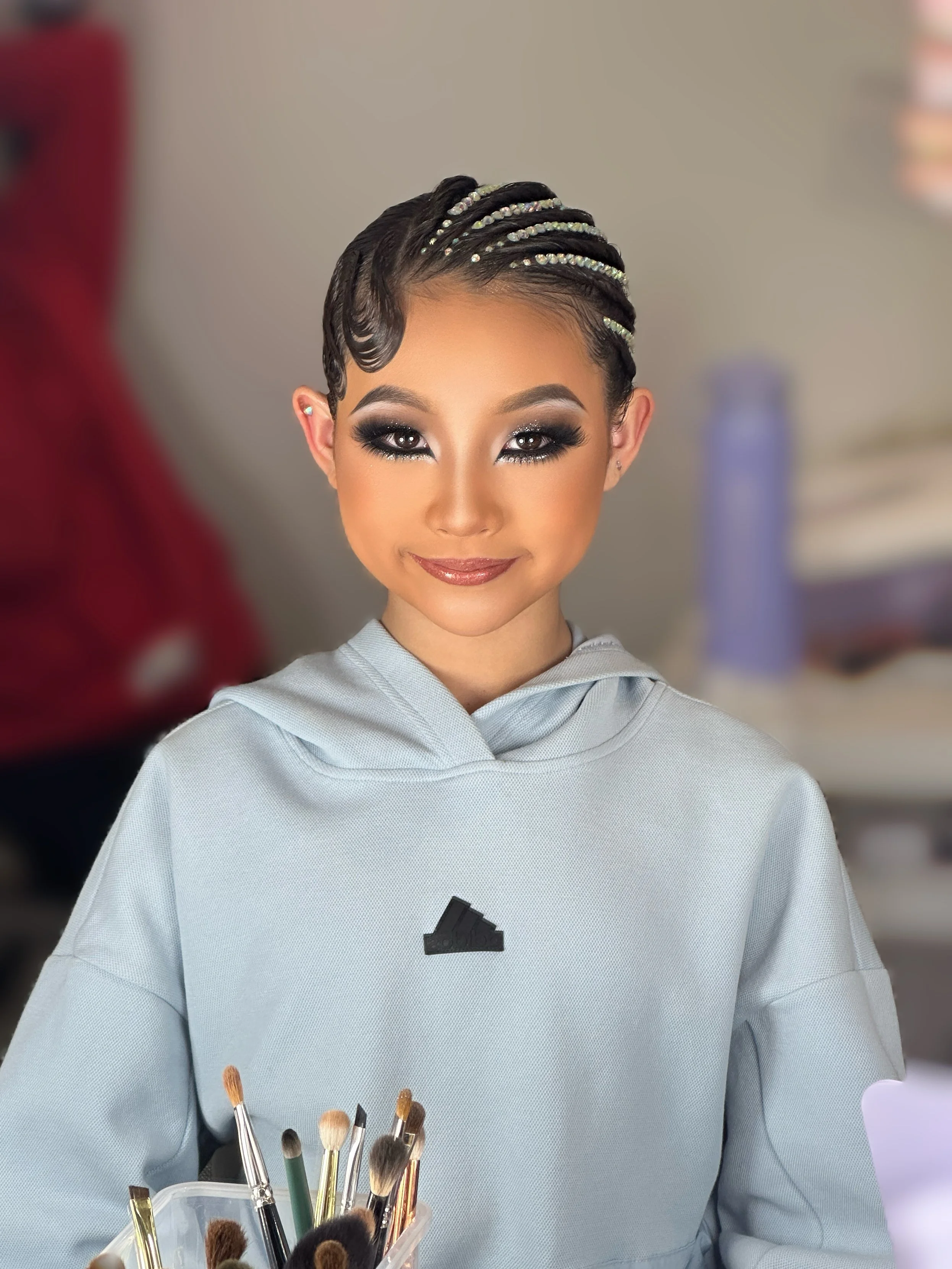 Young woman with styled hair and makeup, wearing a light gray hoodie with a small black logo, holding a container of makeup brushes, smiling at the camera.