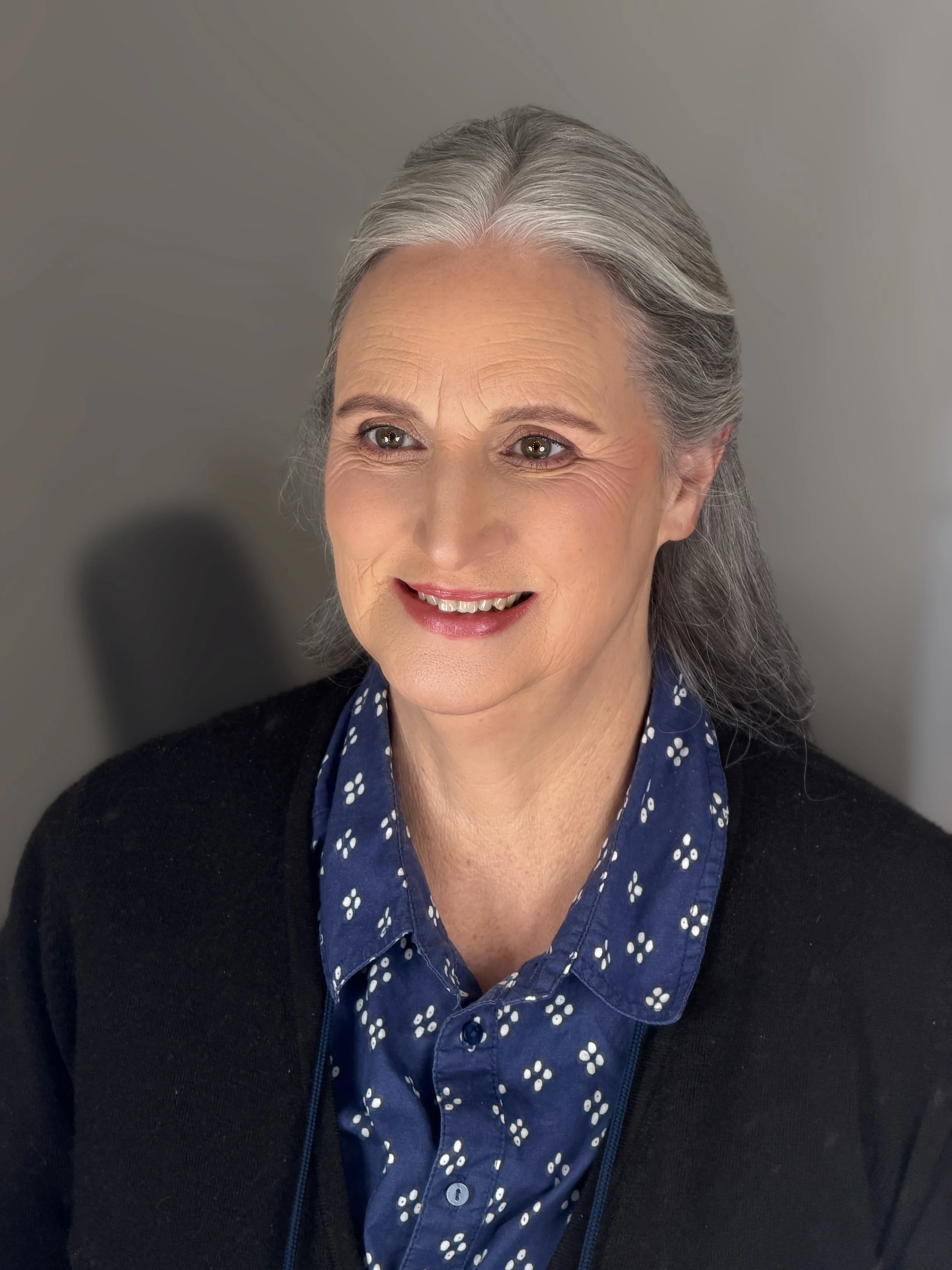 Close-up of a smiling mature woman with gray hair tied back, wearing a blue shirt with white floral pattern and a black sweater, against a neutral background.