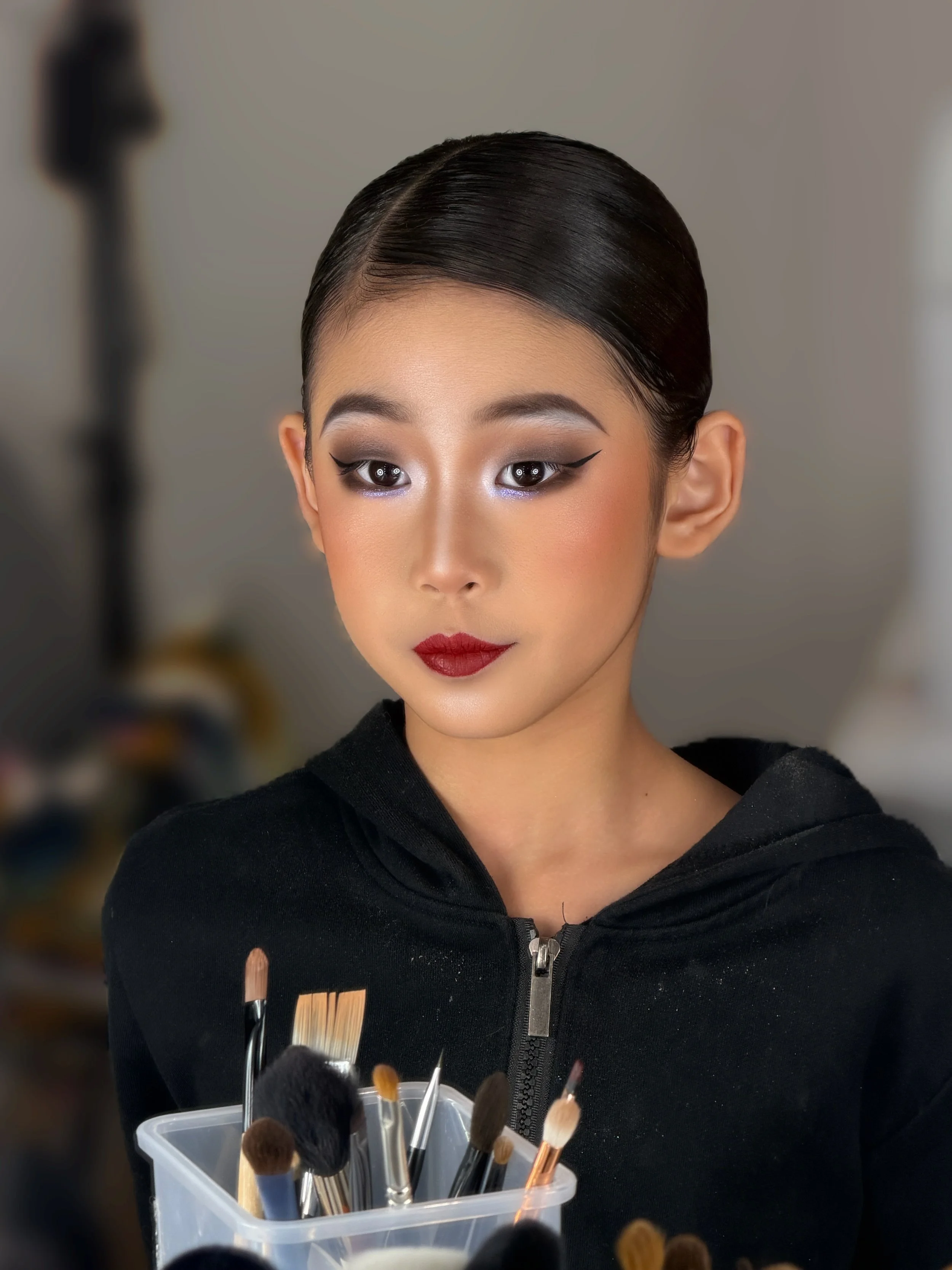 A woman with styled dark hair and makeup, wearing a black zip-up garment, looks into a camera at a makeup station with brushes in a container in the foreground.