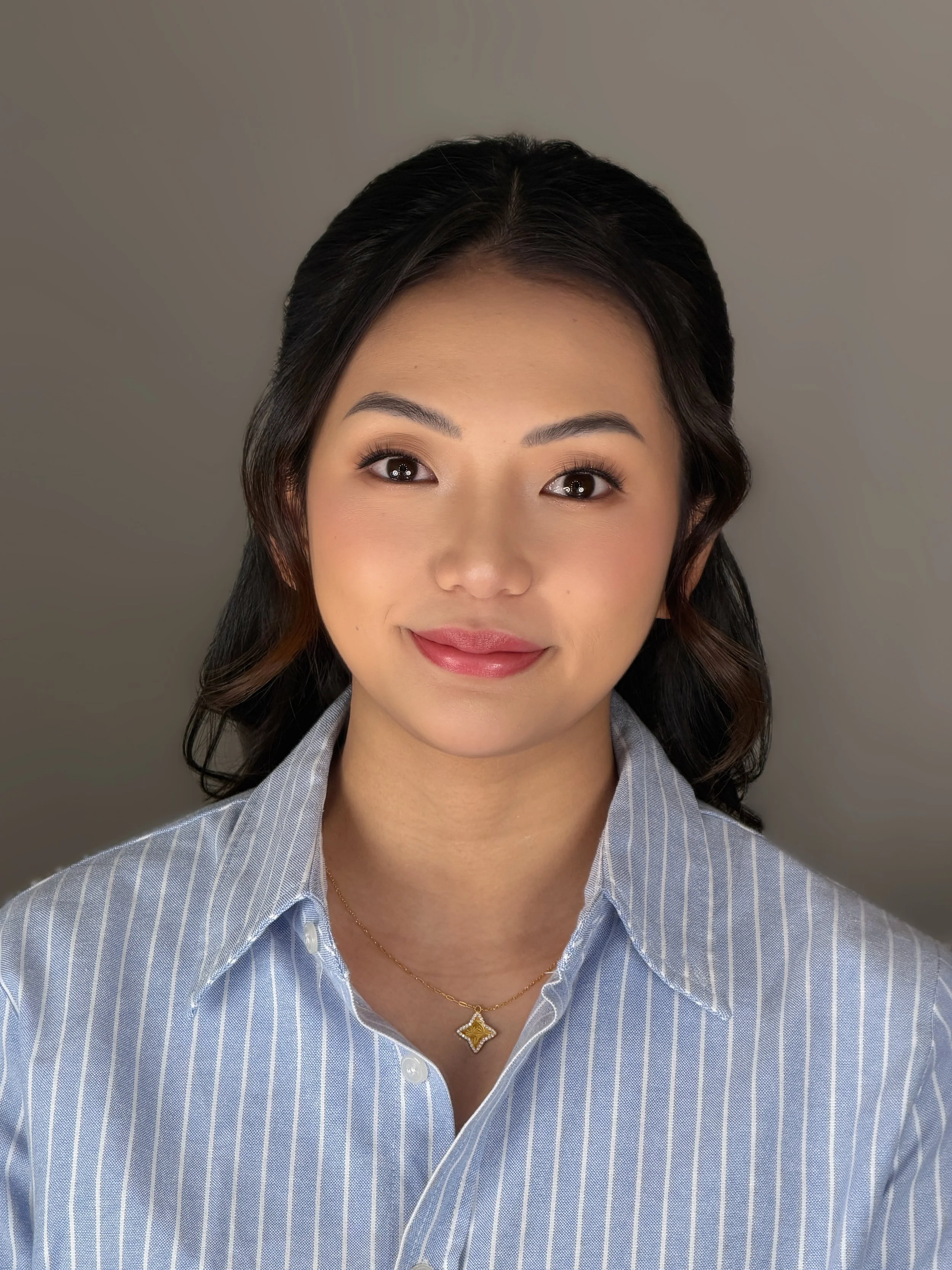 Close-up of a woman with dark hair, smiling, wearing a striped blue and white shirt, gold necklace, and a pendant, against a plain background.
