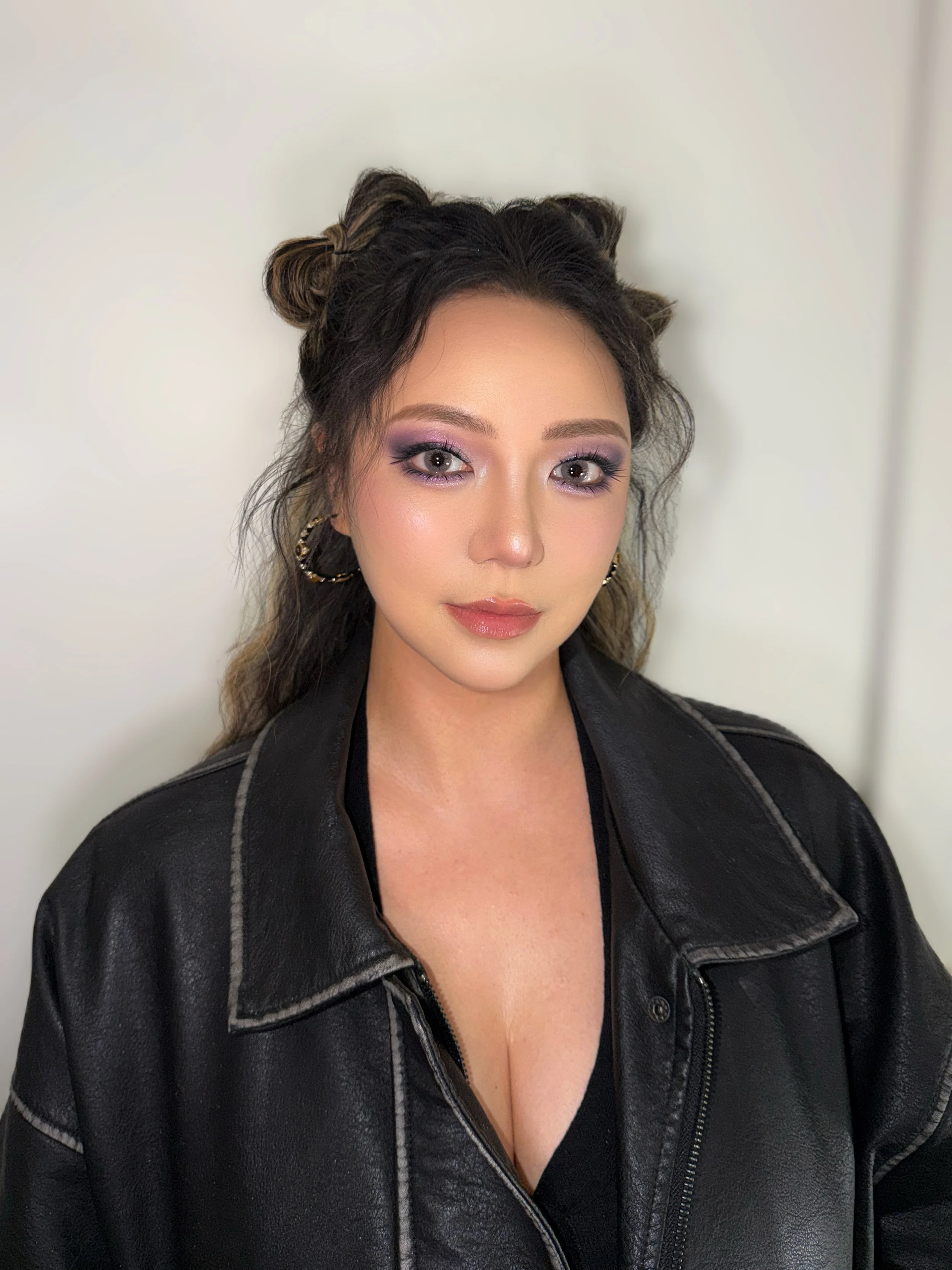 Close-up of a woman with styled hair, wearing makeup, gold hoop earrings, and a black leather jacket.