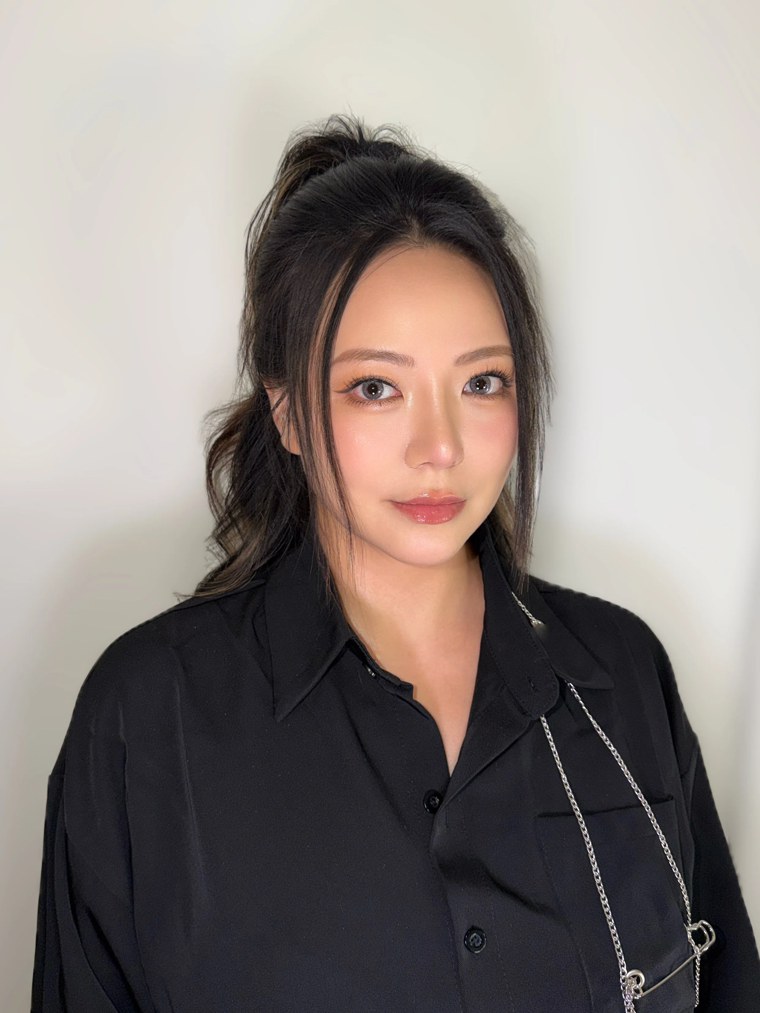 A woman with black hair styled in a half-up, half-down look, wearing a black button-up shirt and a silver chain necklace, standing in front of a plain white wall.