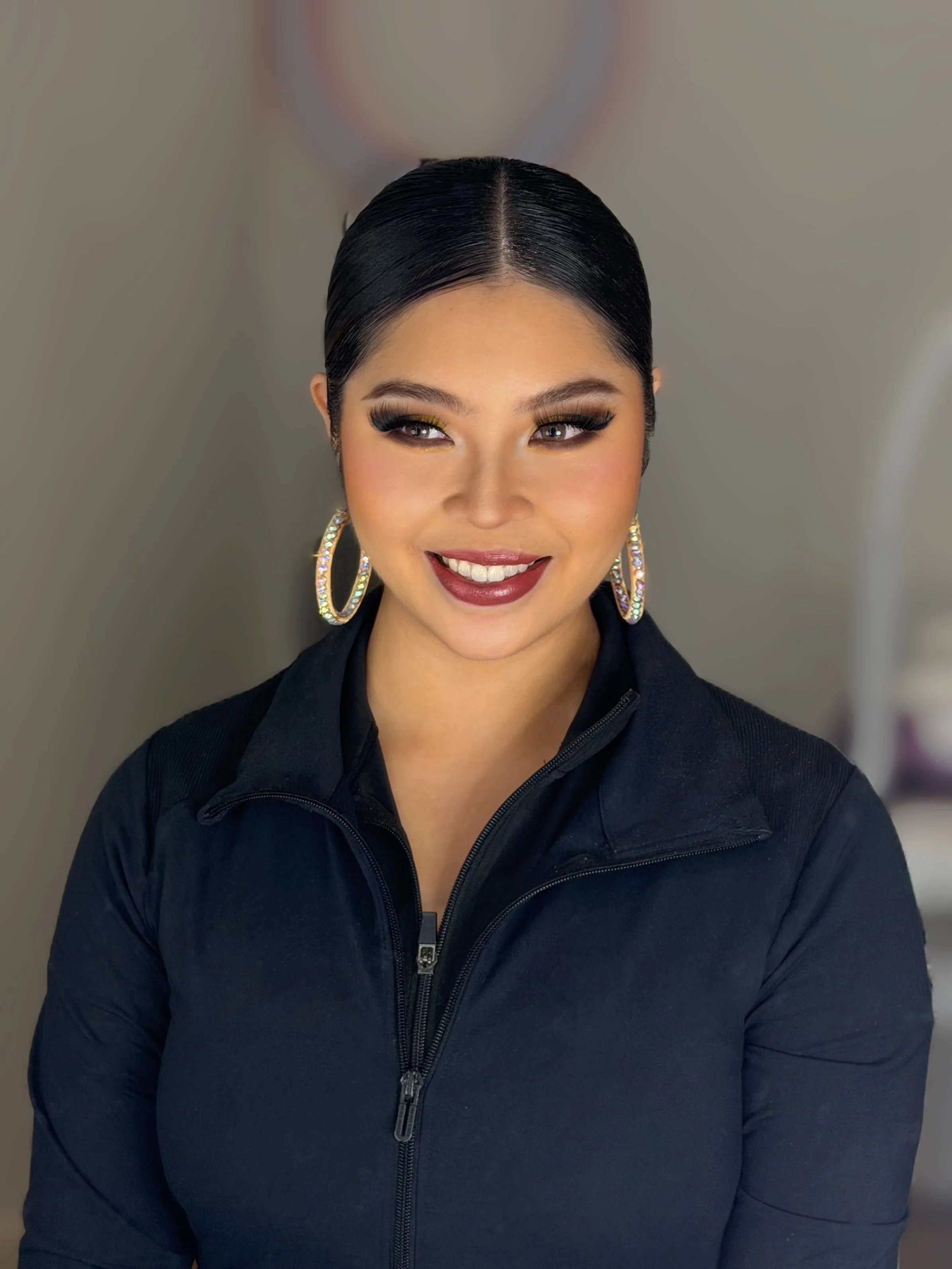 A woman with dark hair slicked back, wearing large hoop earrings with rhinestones, makeup with bold eye makeup and lipstick, smiling, dressed in a black zip-up jacket.