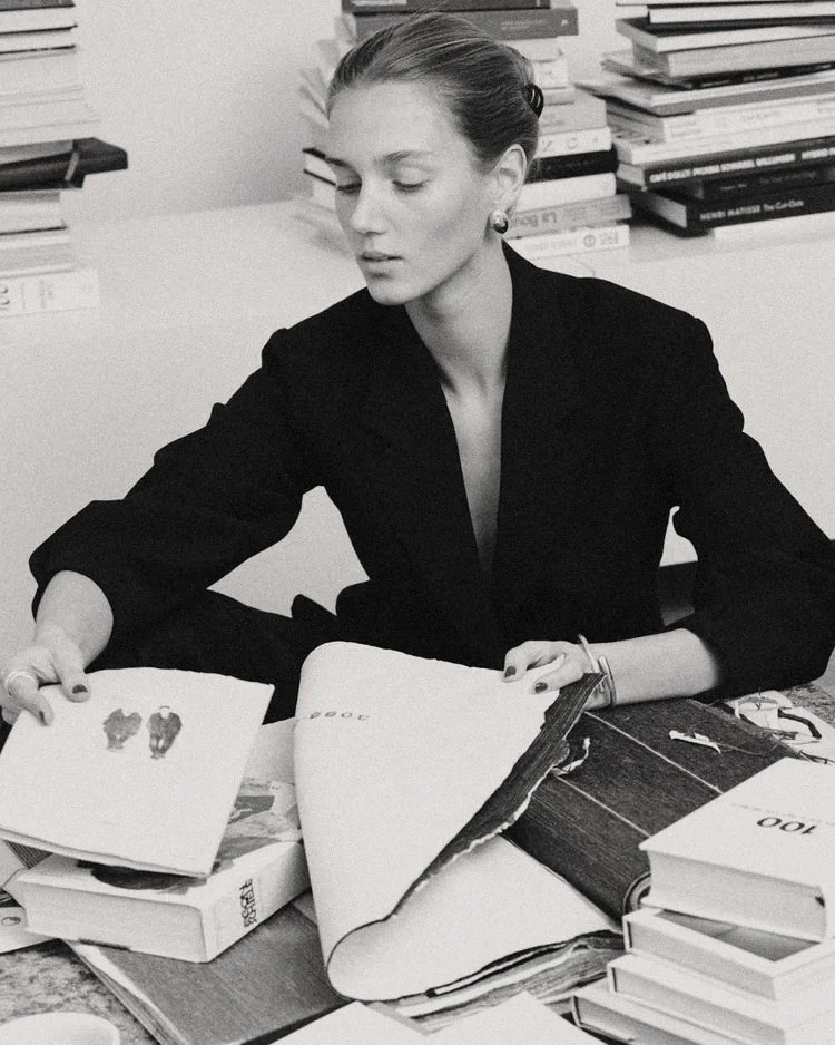 A woman with hair in a bun, dressed in a black blazer, sitting at a cluttered desk with books and papers, looking down at an open book.