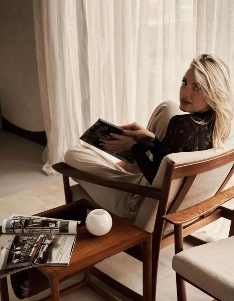 A woman with blonde hair sitting in a beige armchair, holding a magazine, in a cozy room with light-colored curtains. A wooden coffee table in front holds an open magazine and a small white decorative object.