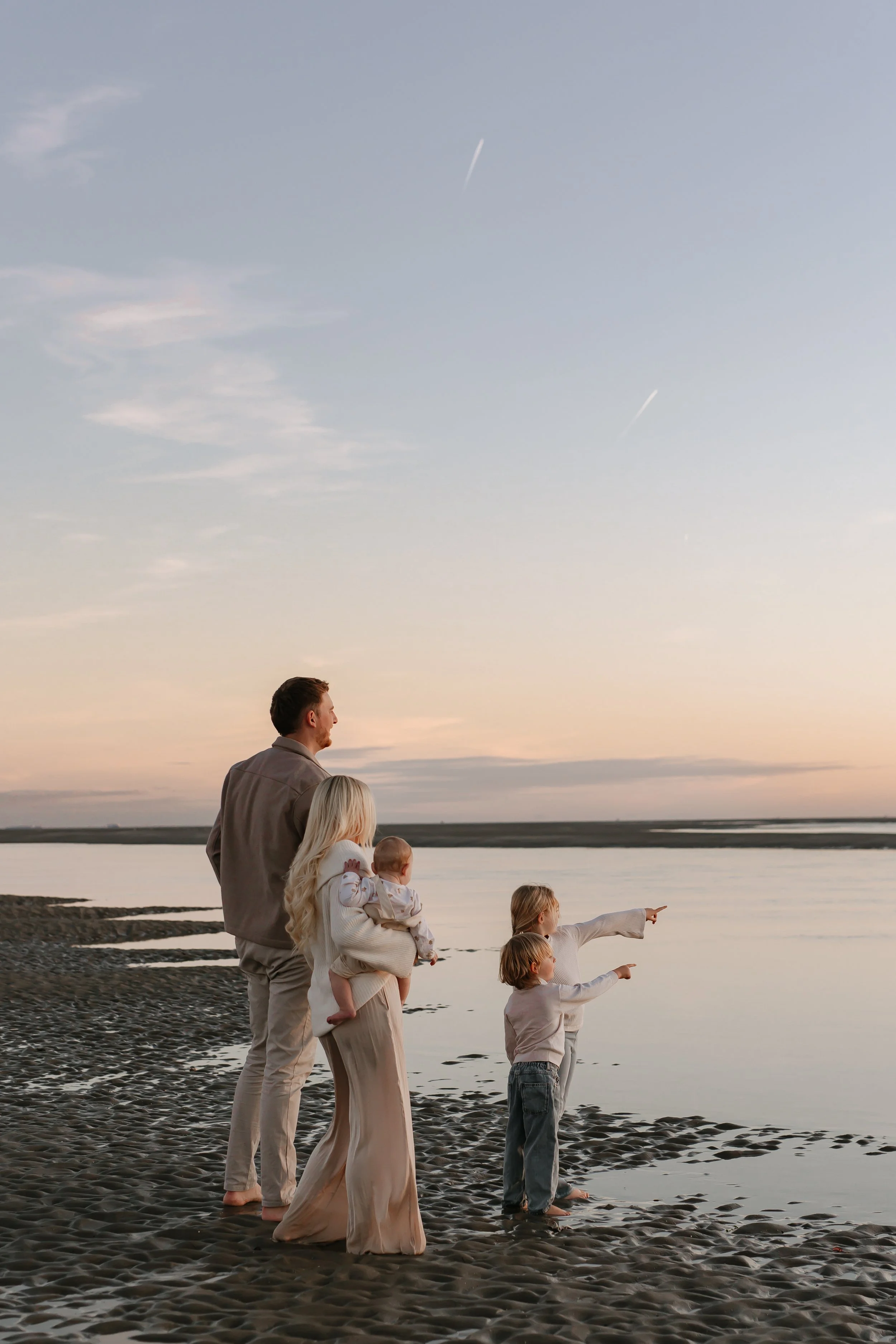 A family of five, including two adults and three children, standing on a beach at sunset, looking out at the water and pointing towards the sky.