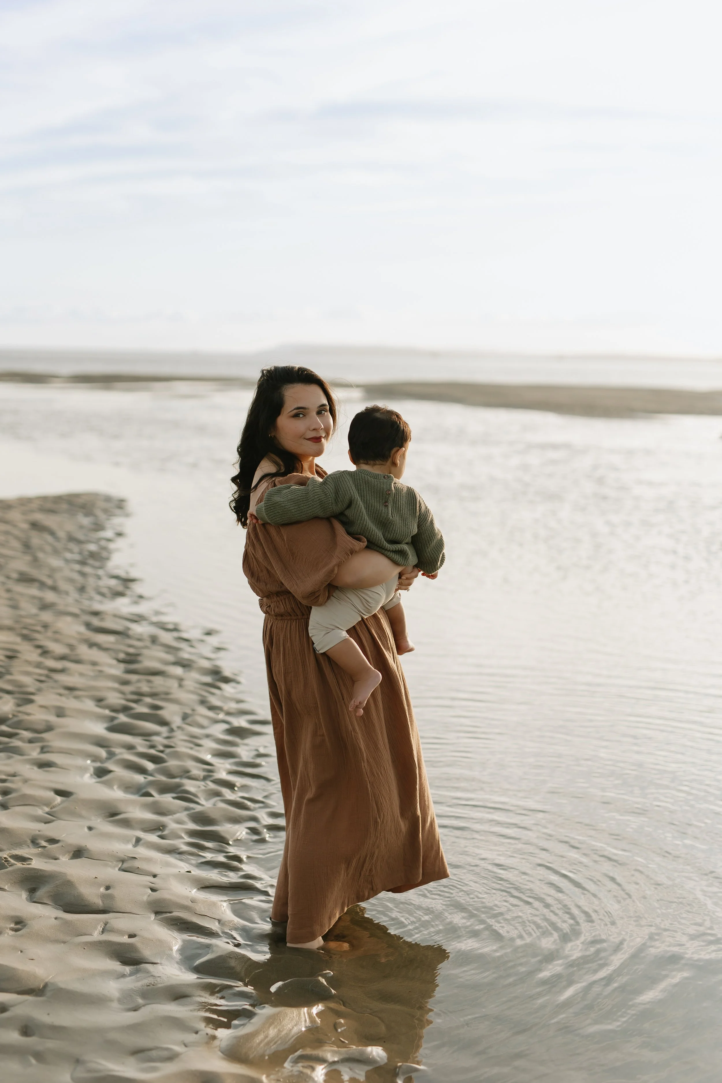 A woman in a long brown dress holding a child in a green sweater on a sandy beach by the water at sunset.