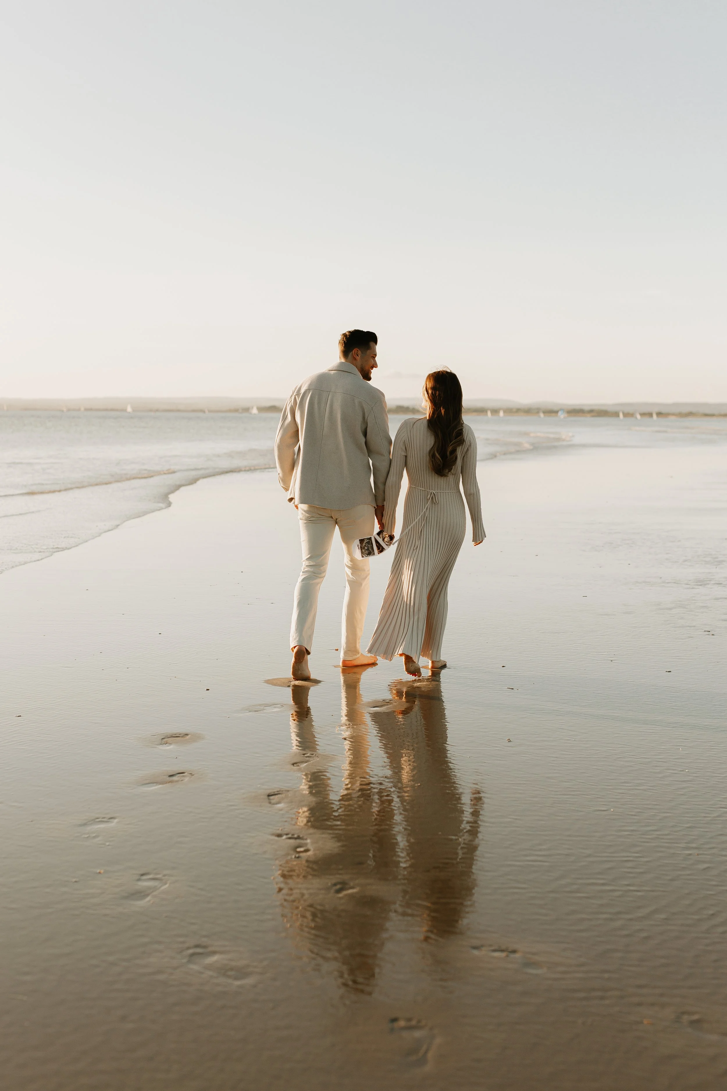 A couple holding hands and walking barefoot on the beach during sunset, with gentle waves and sailboats in the distance.