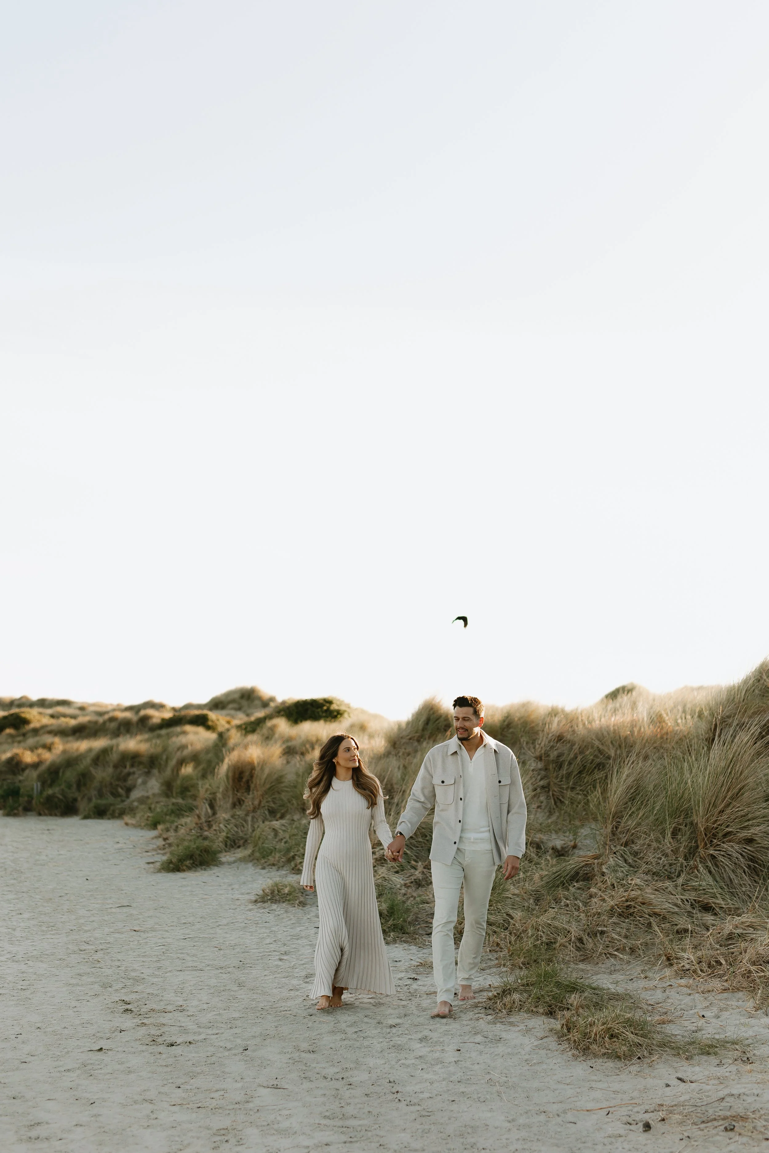 A couple walking hand in hand on a sandy beach with grassy dunes, under a clear sky, appearing happy and relaxed.
