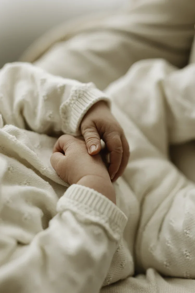 A close-up of an adult holding a baby's hand, both wearing cream-colored sweaters.