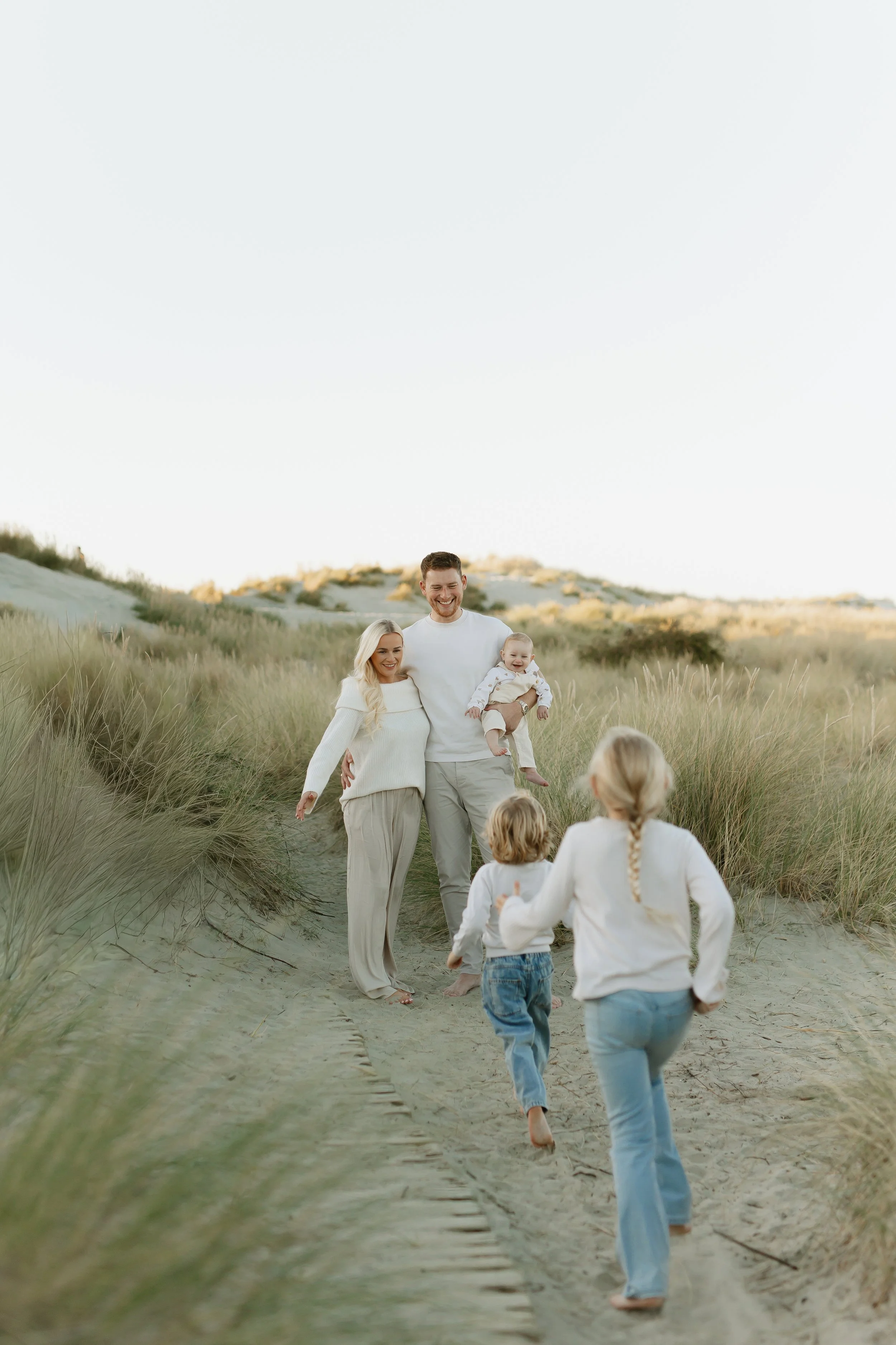 A happy family walking on a sandy path through dunes with tall grass, with a clear sky overhead.