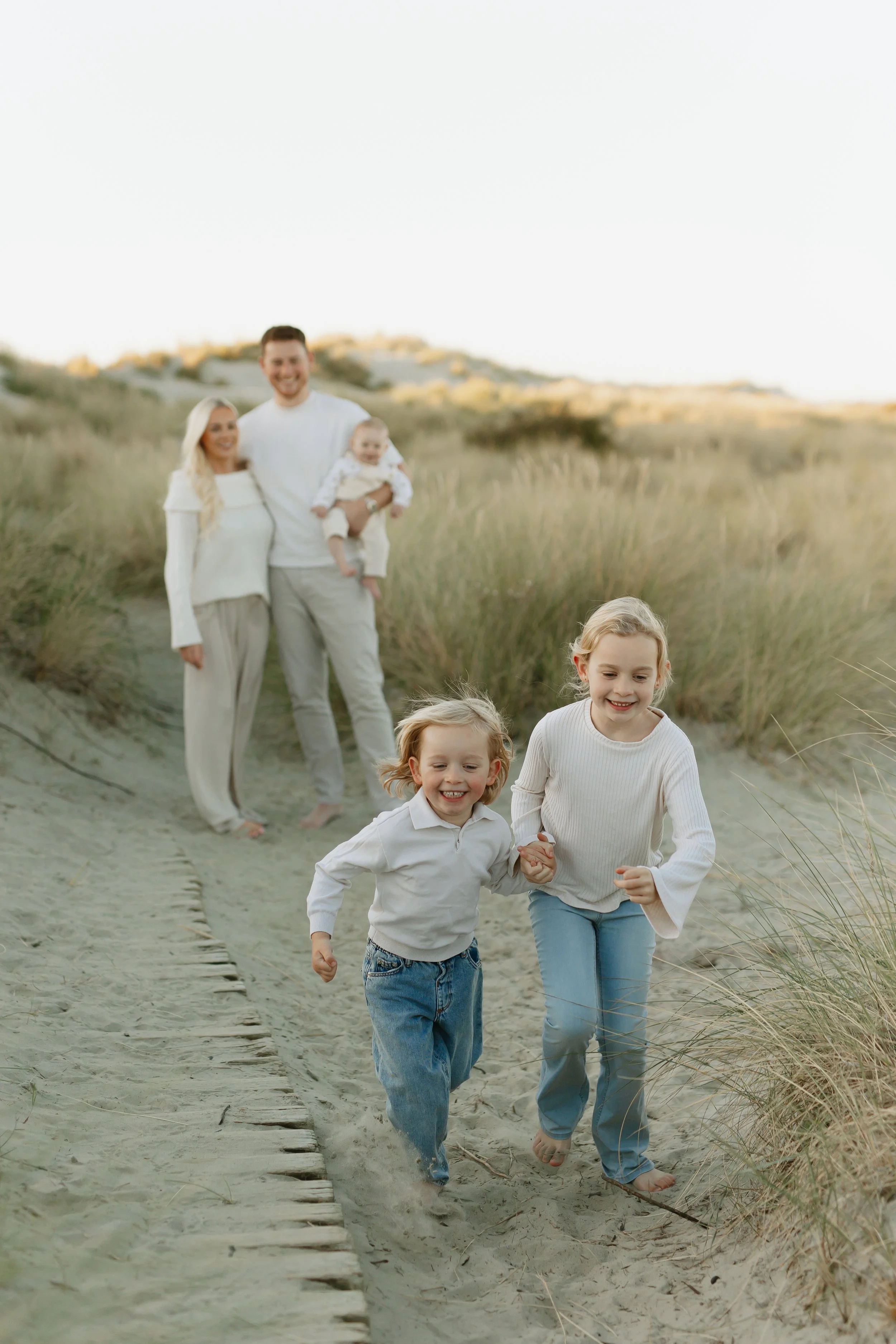 A family of four running on a sandy beach with grassy dunes in the background. The children are at the front, holding hands, smiling and barefoot. The parents walk behind, smiling, with the father holding a baby.