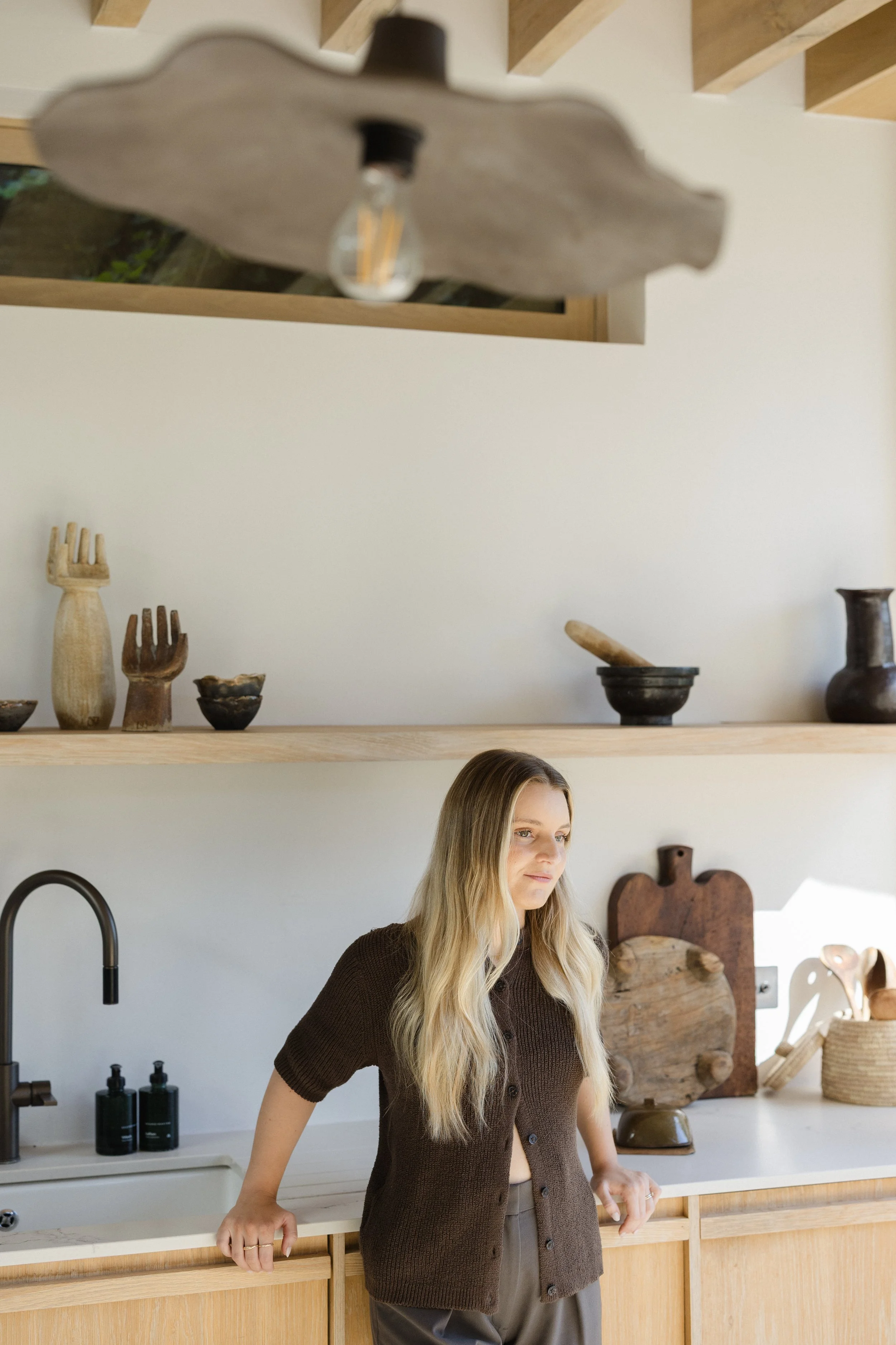 A woman with long blonde hair wearing a brown knitted cardigan and gray pants standing in a modern kitchen with wooden cabinets, black faucet, and decorative pottery on shelves.