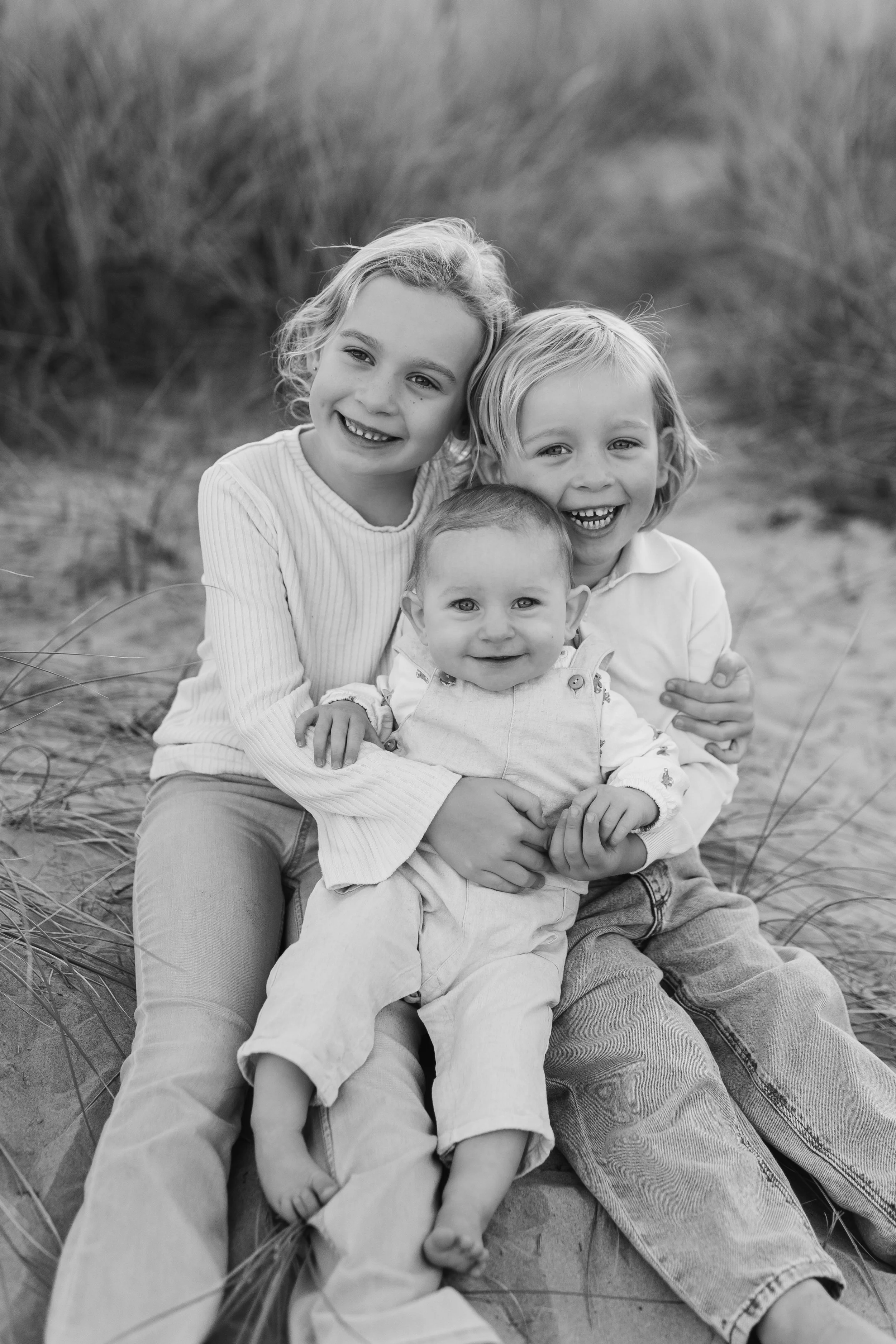 A black and white photo of three young children sitting on sand with tall grass in the background. They are smiling and hugging each other.