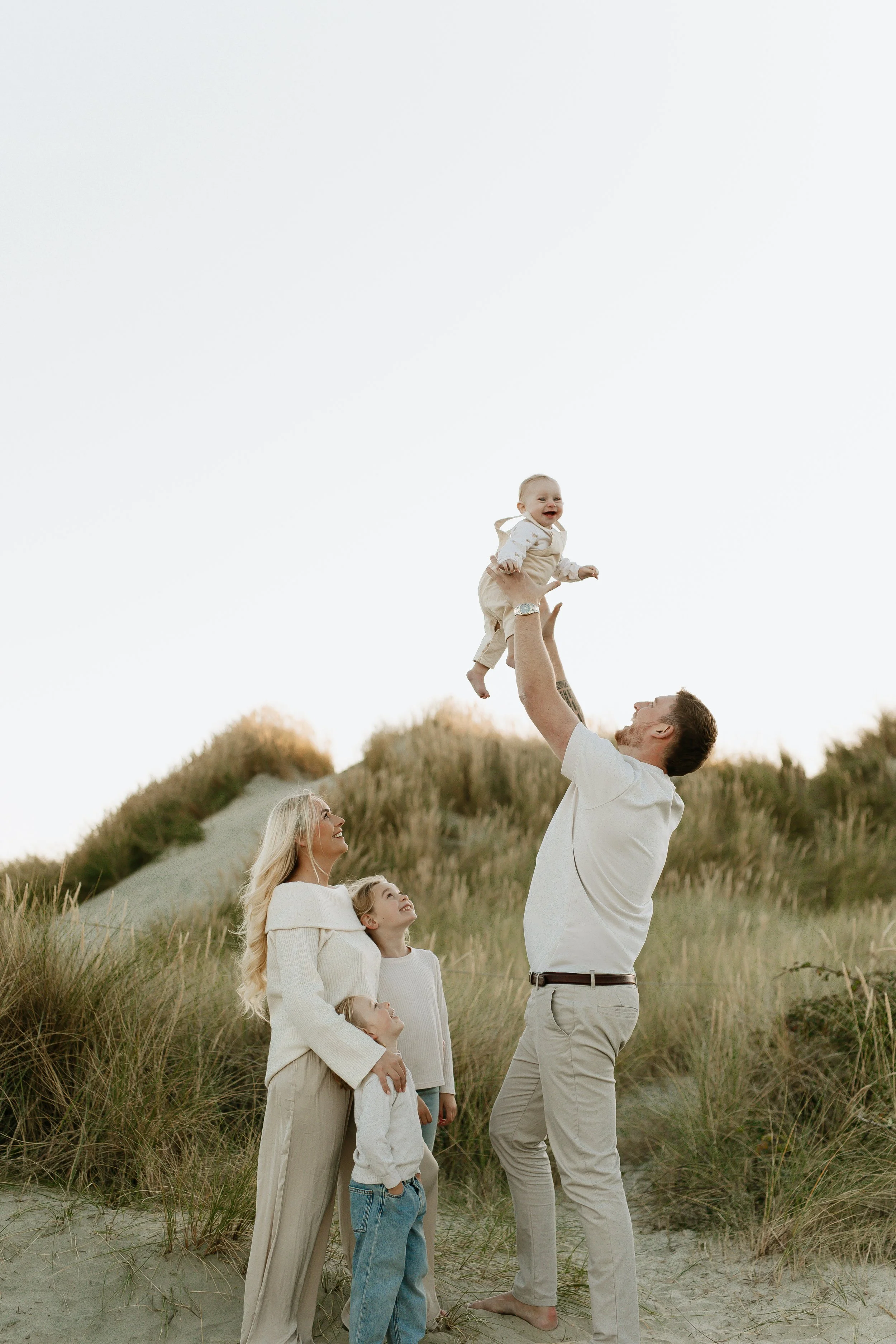 Family of four outdoors in a sandy area with dunes and tall grass, a man lifts a smiling baby in the air while a woman and two children look on happily.