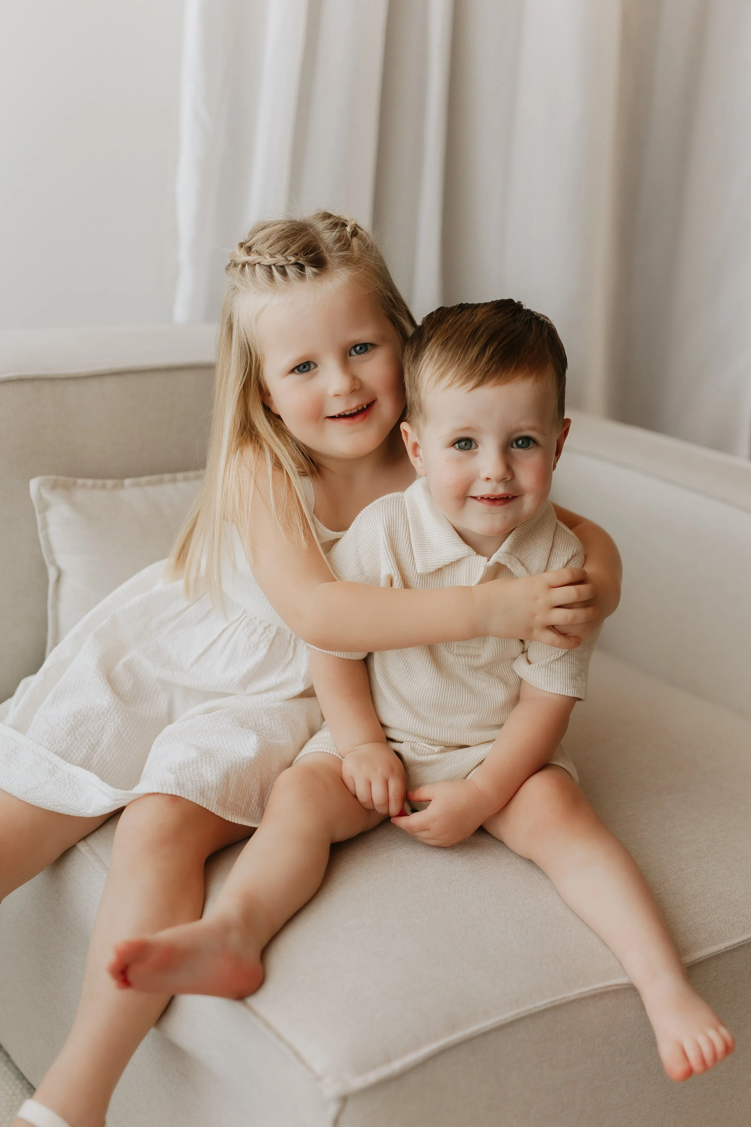 Two young children, a girl and a boy, sitting on a beige sofa, smiling, with the girl hugging the boy from behind, in a bright room with white curtains.