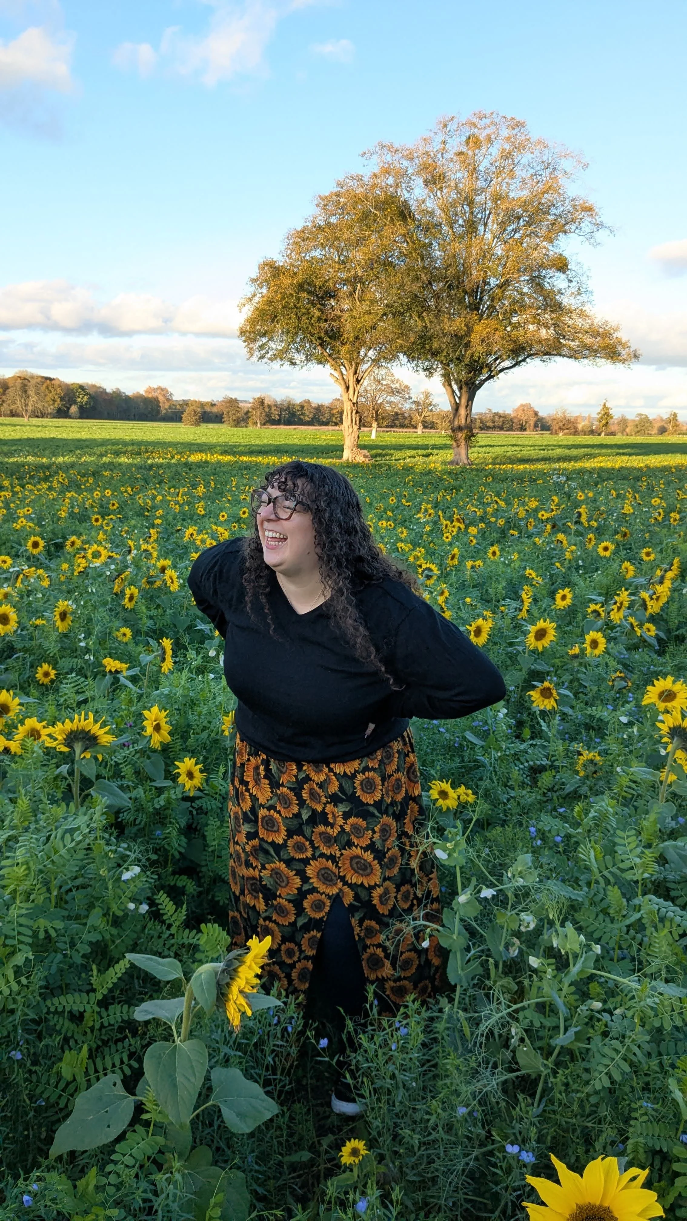 A woman with curly black hair, glasses, and a black top stands in a sunflower field, smiling and laughing with hands behind her back. Large trees are in the background under a blue sky with scattered clouds.