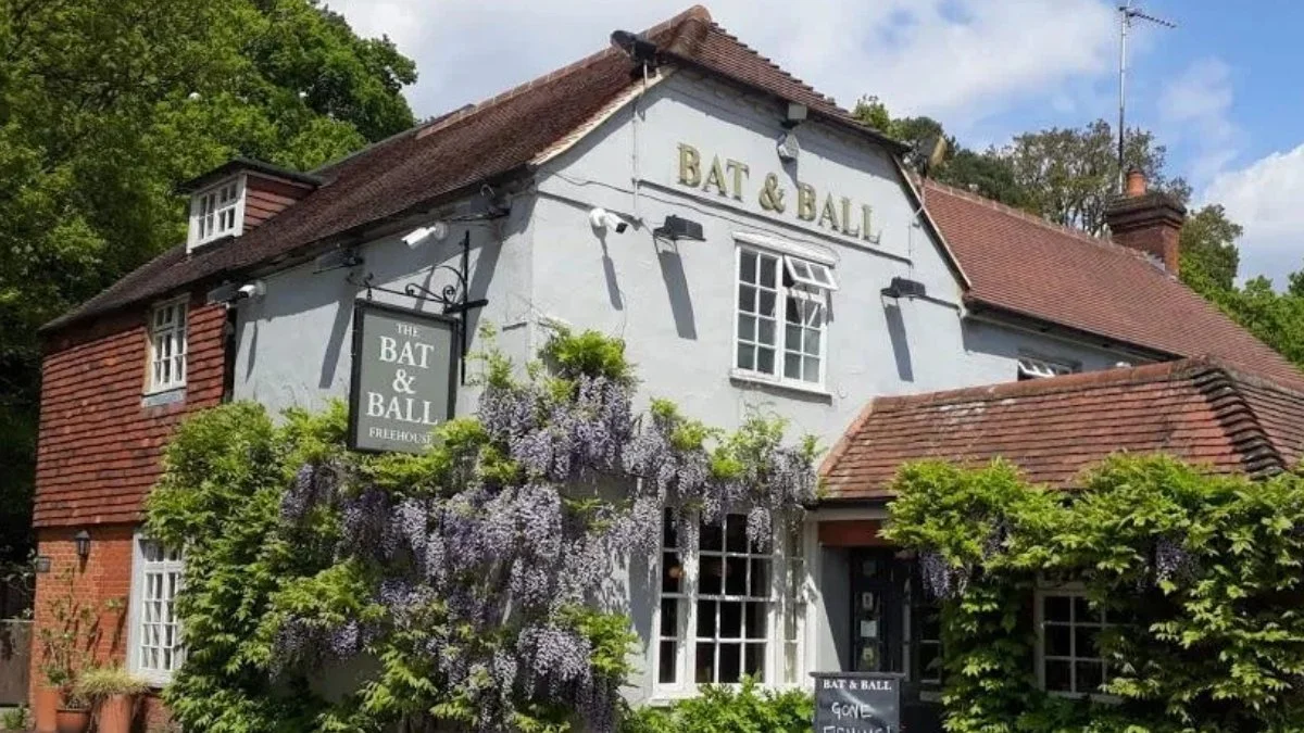 The white building with a red tiled roof has a sign for "The Bat & Ball" pub. There is greenery and purple flowering vines in front, and a small chalkboard sign near the entrance.