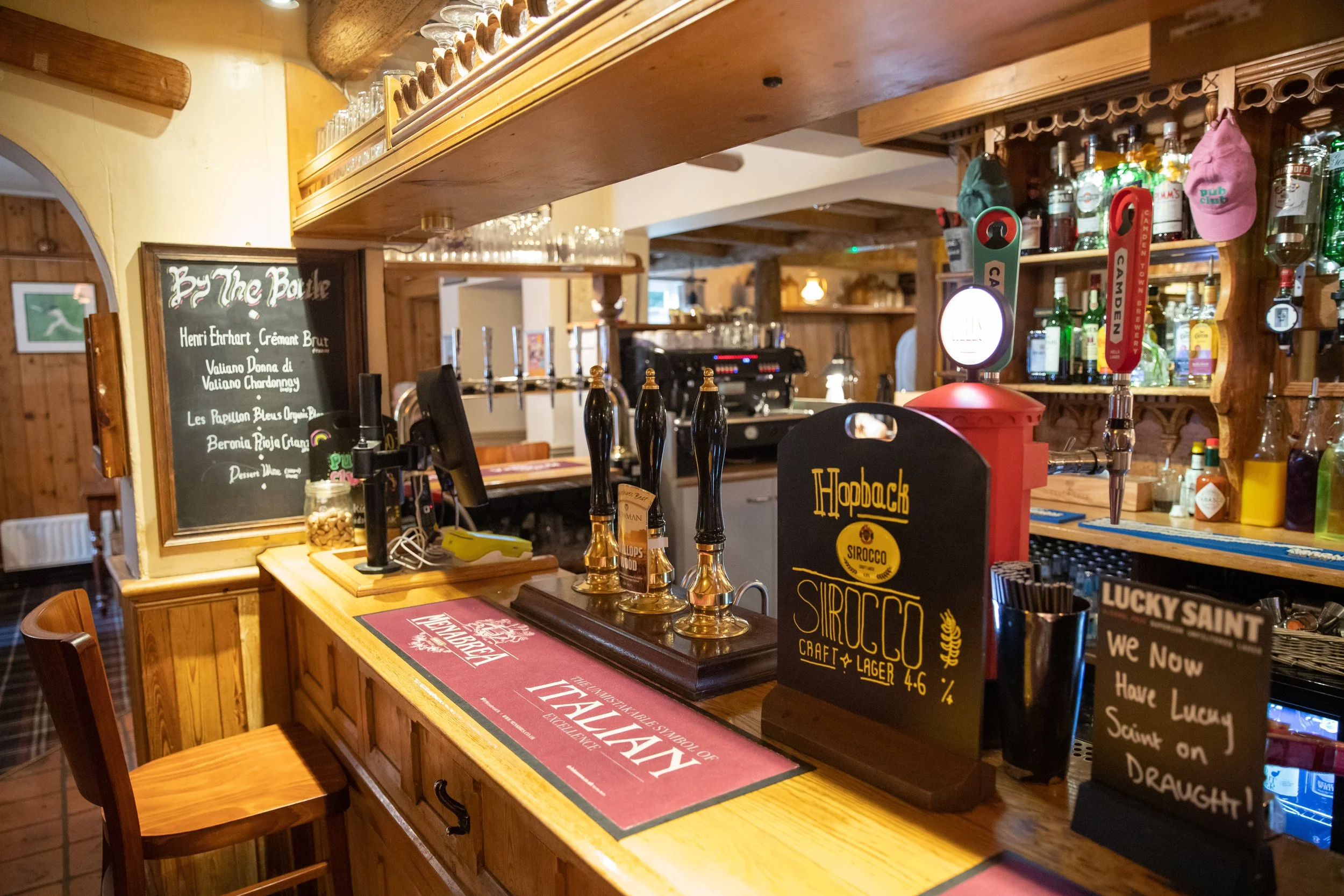 Interior of a cozy bar with a wooden counter, beer taps, shelves with liquor bottles, and chalkboard signs displaying drink options.