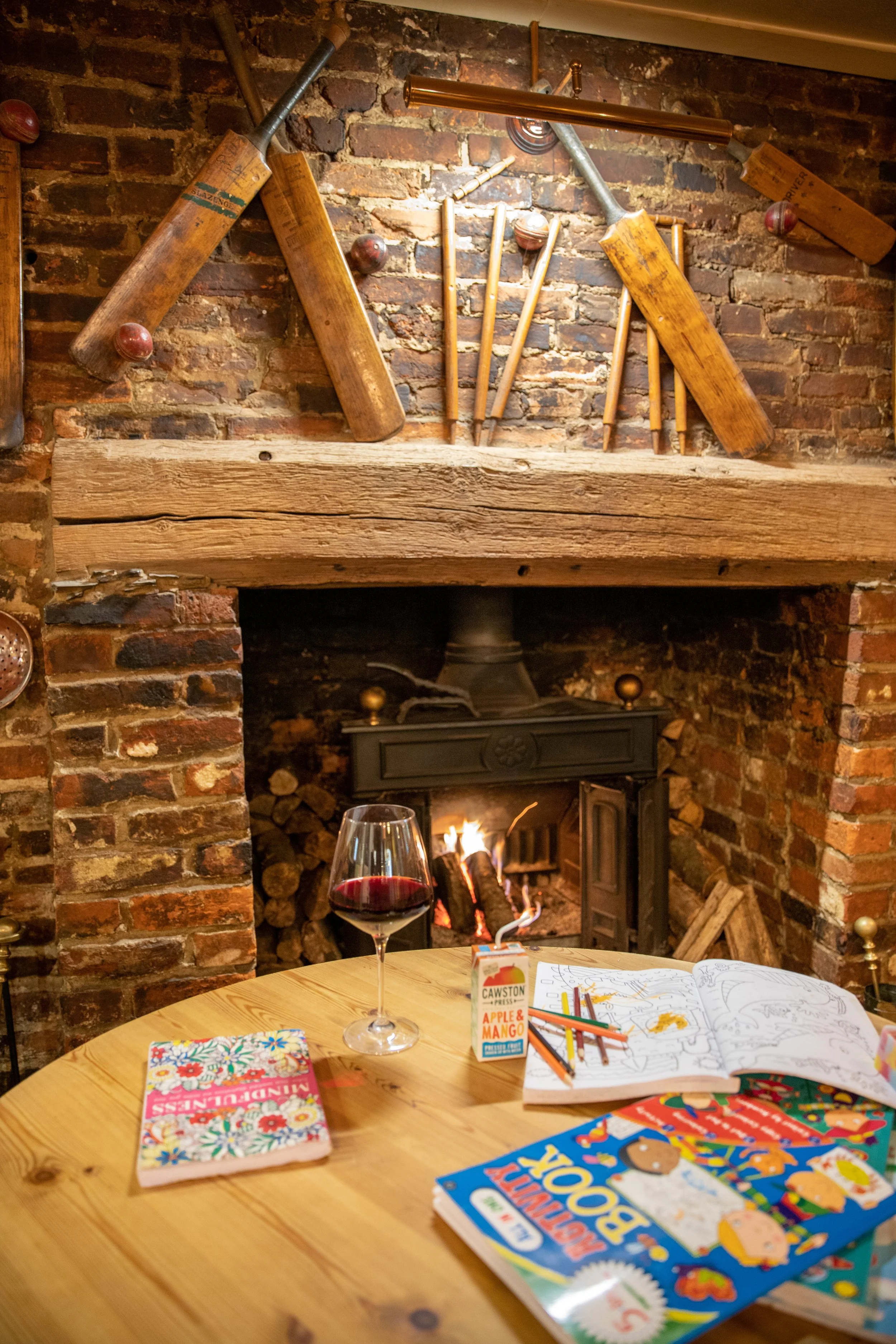 A cozy indoor scene with a round wooden table holding a glass of red wine, crayons, coloring pages, a juice box, and a book. In the background, a brick fireplace with stacked logs and a fire burning, above which a rustic brick wall displays vintage w