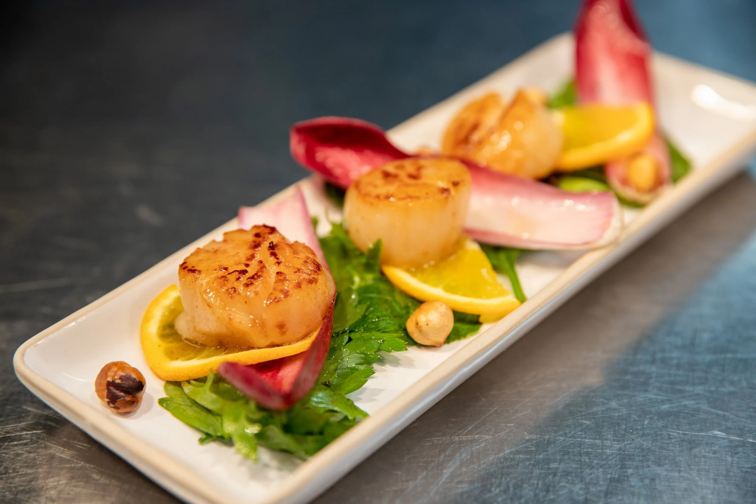 Seared scallops served on a white rectangular plate with lemon slices, radish, greens, and nuts, presented in a fine dining style.