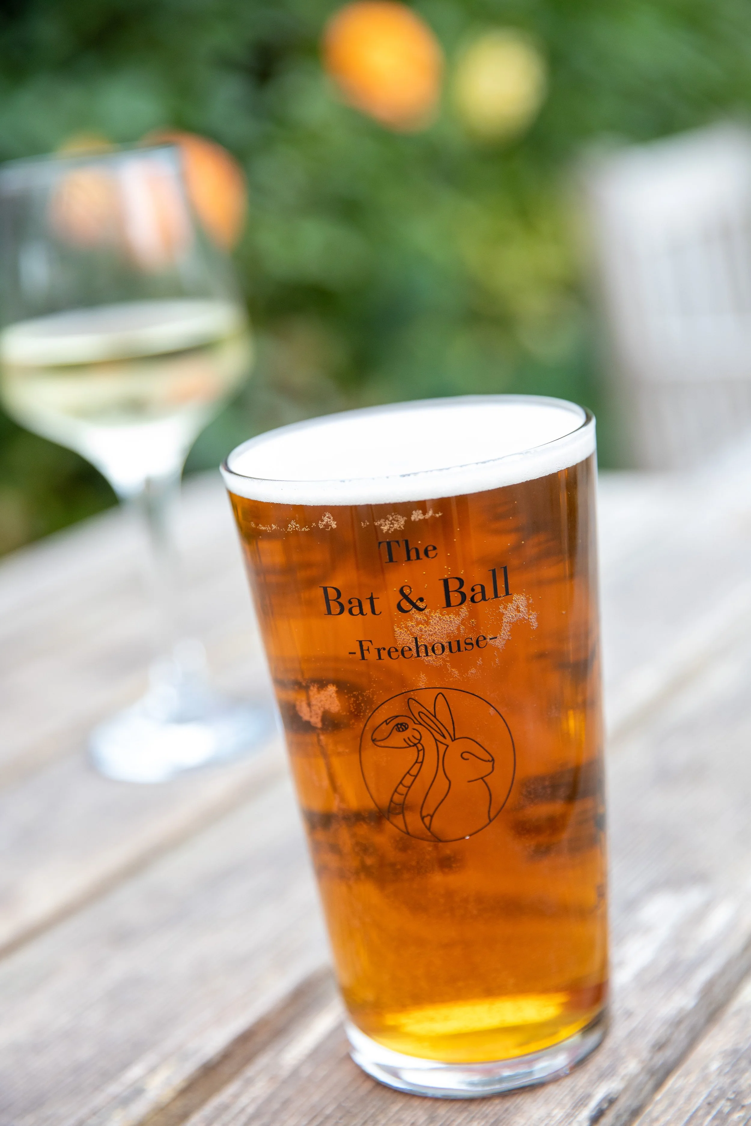A glass of beer with a logo that reads 'The Bat & Ball - Freehouse' and an illustration of a rabbit and a snake. In the background, there is a glass of white wine on a wooden table.