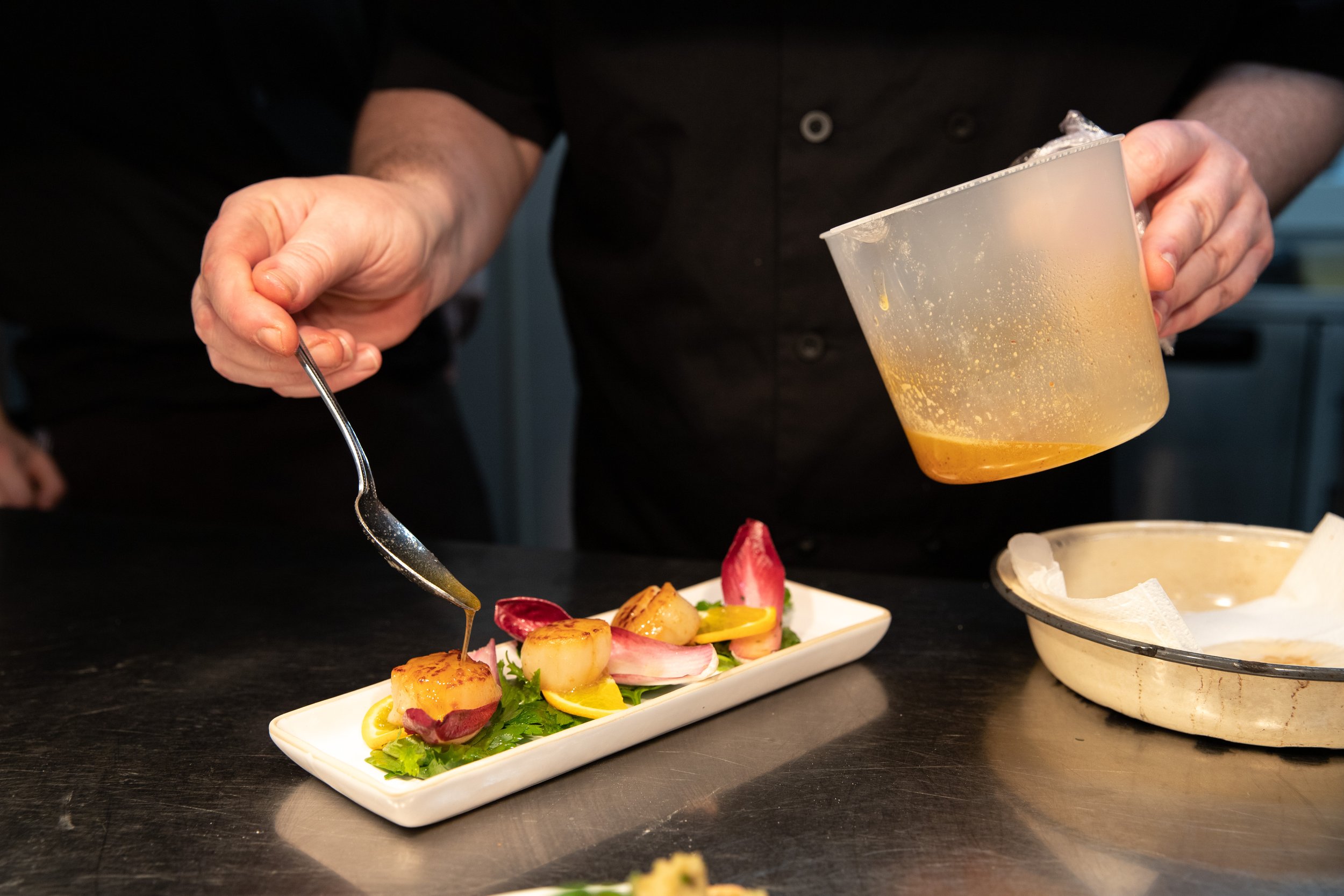 Chef drizzling sauce over seared scallops garnished with greens and lemon slices on a rectangular white plate.