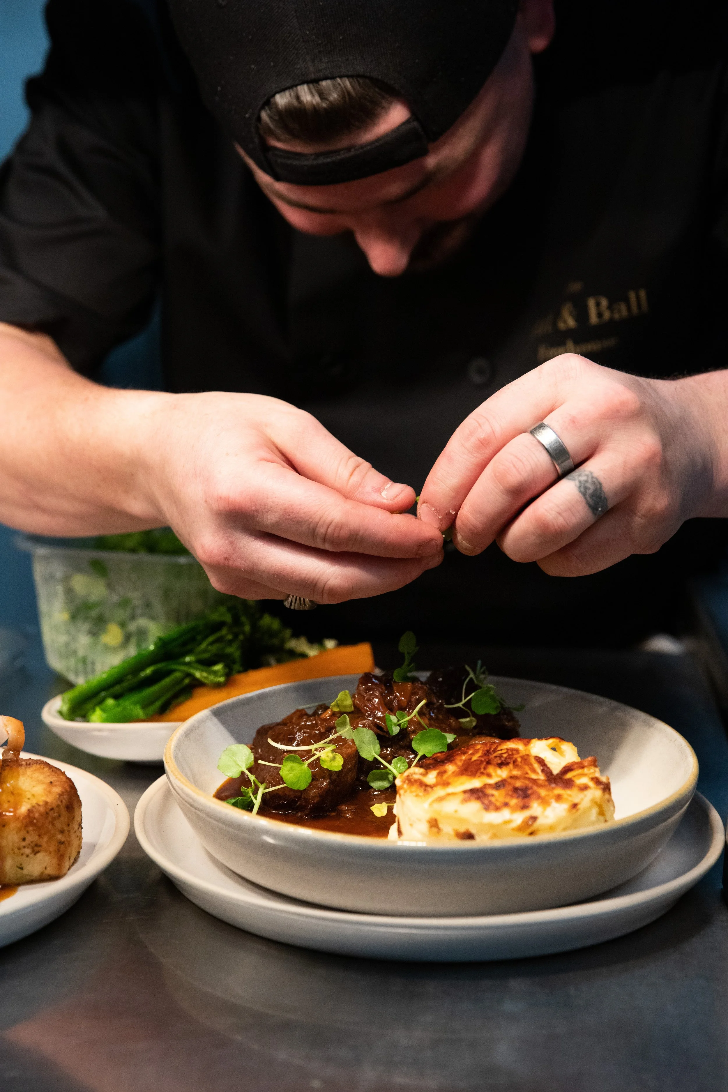Chef garnishing a plated meal with herbs, with side dishes and vegetables on the table.