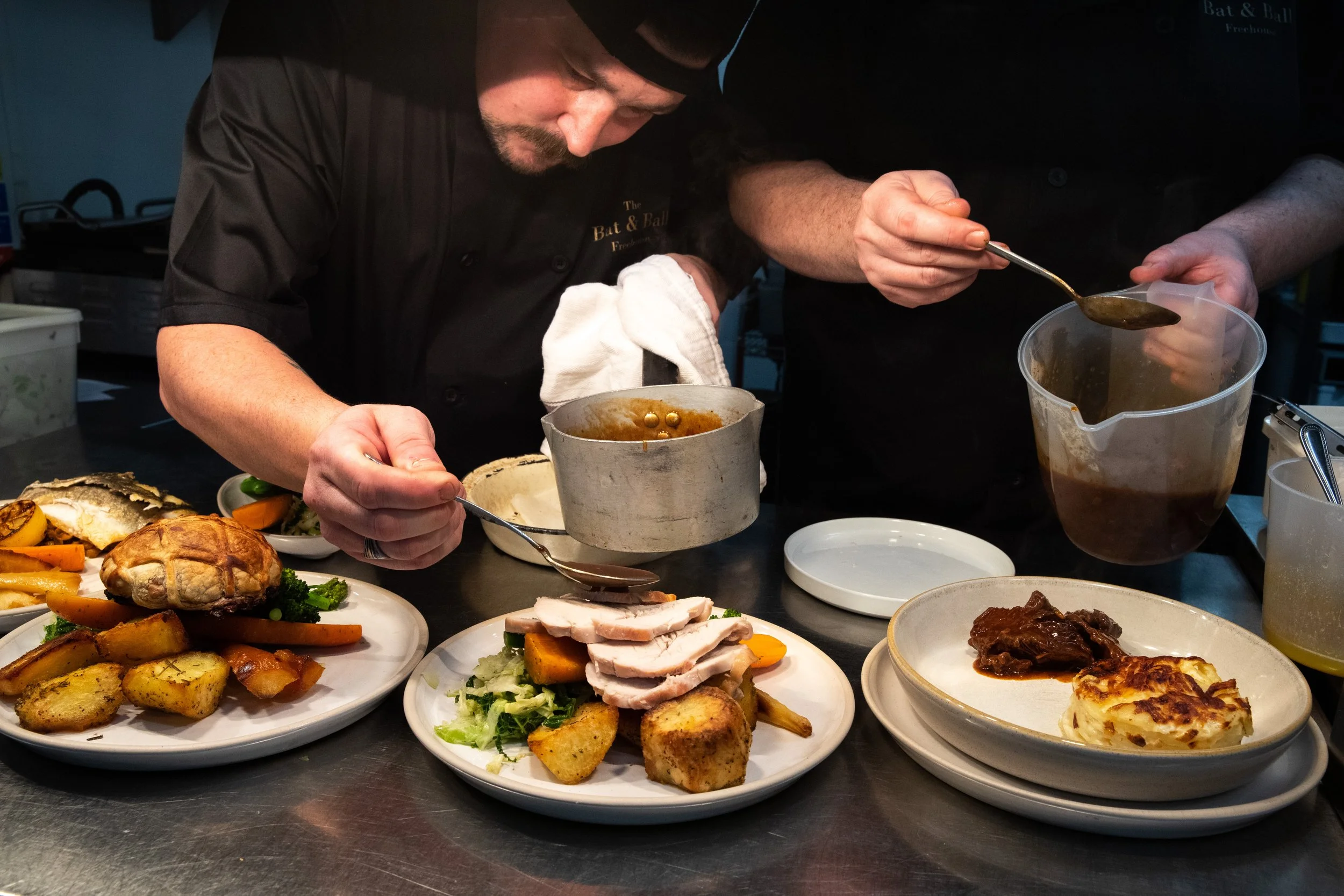 Two chefs preparing plated dishes in a professional kitchen, with various plated foods including roasted potatoes, vegetables, sliced meat, and a casserole.
