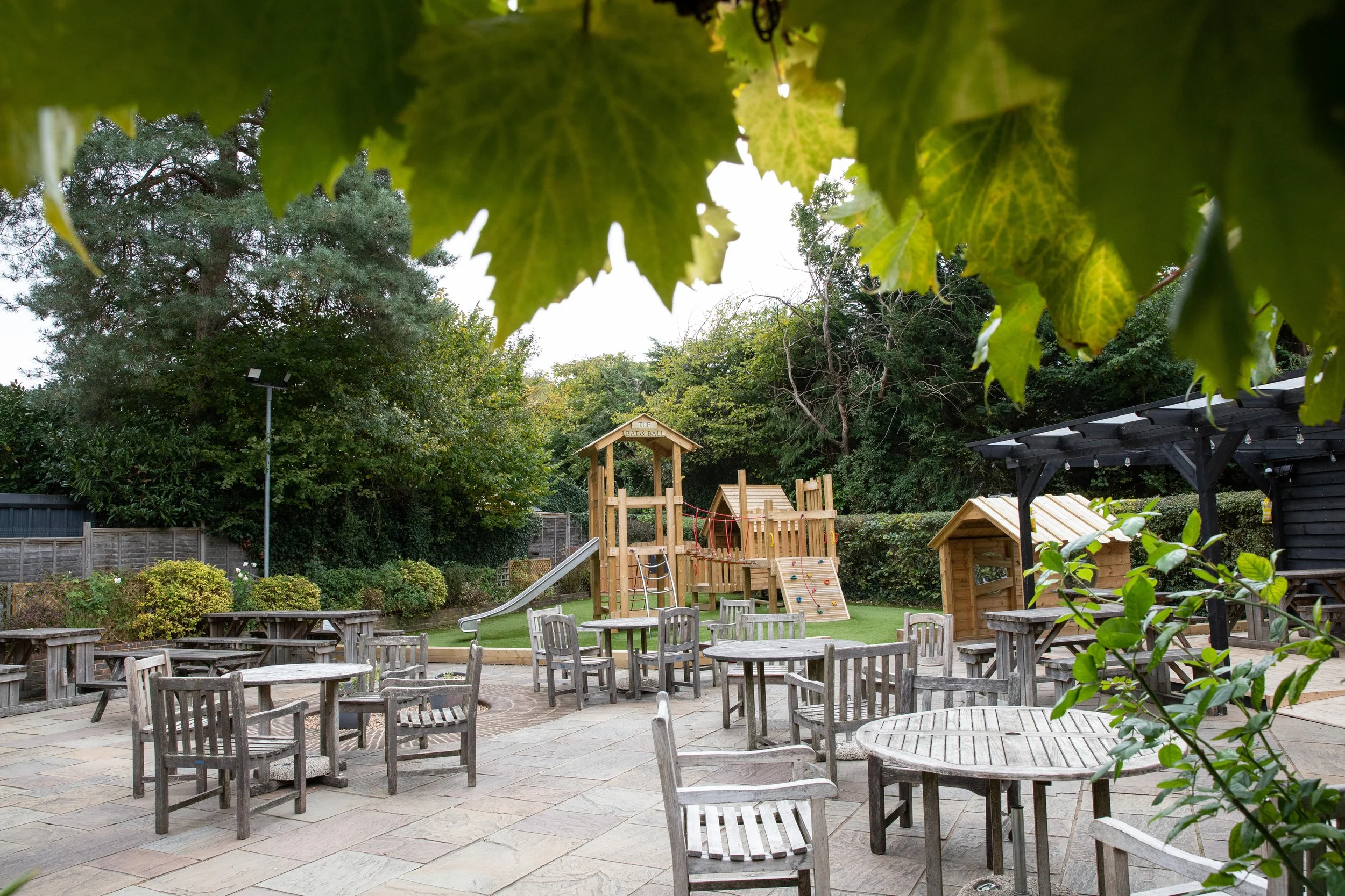 An outdoor patio area with wooden tables and chairs, a playground with a slide and climbing structures in the background, surrounded by trees and greenery.