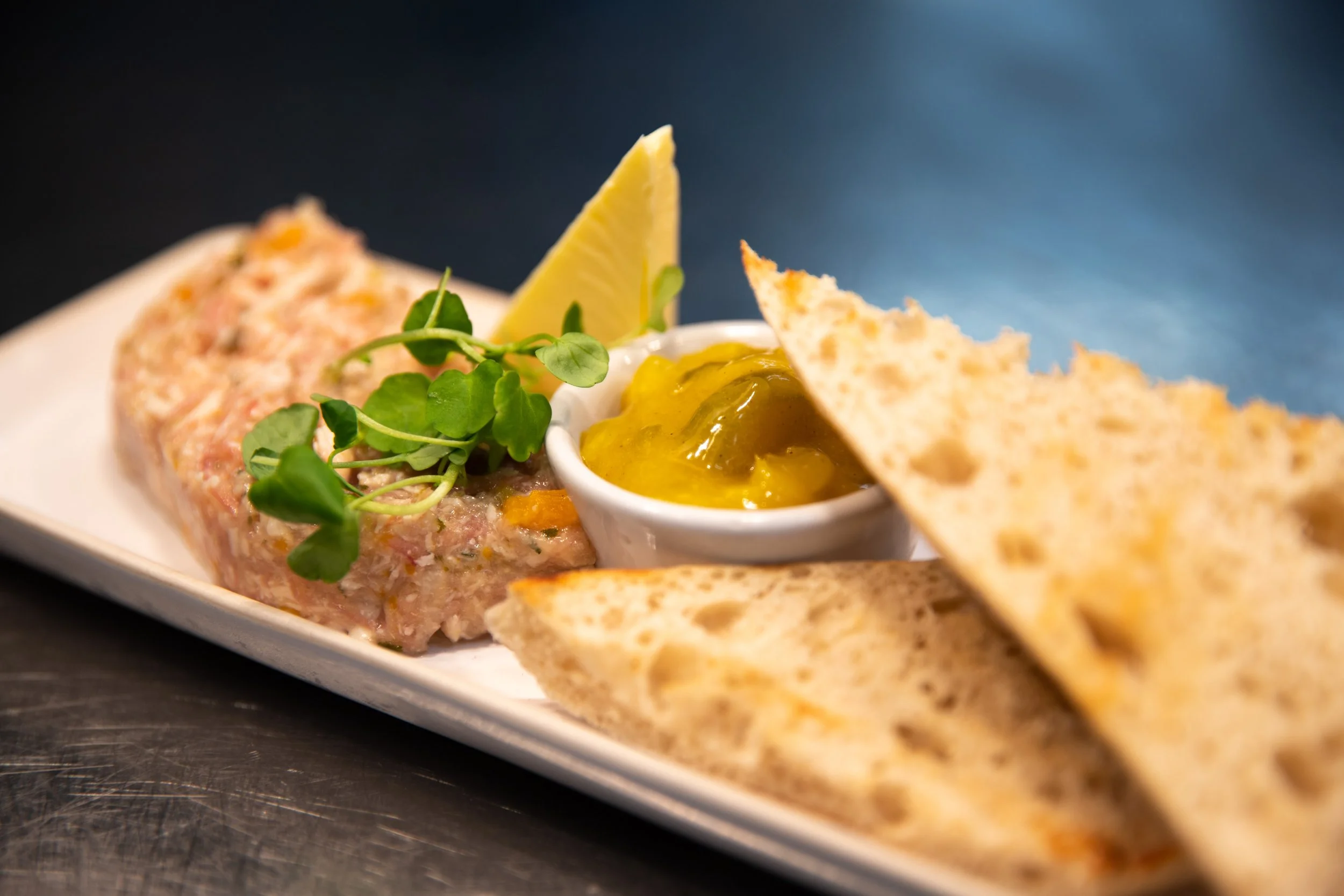 Appetizer plate with tuna tartare garnished with microgreens, a lemon wedge, a ramekin of yellow sauce, and toasted flatbread triangles on a white rectangular dish.