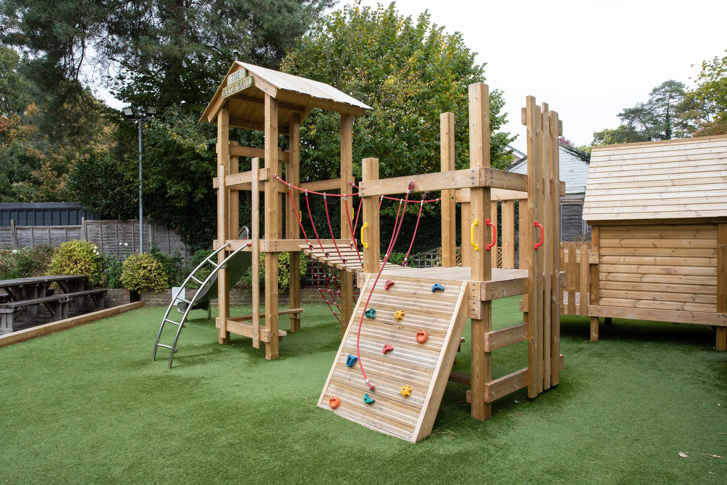 A wooden playground structure with a slide, climbing wall, and ropes, set on a grassy yard with trees and a fence in the background.