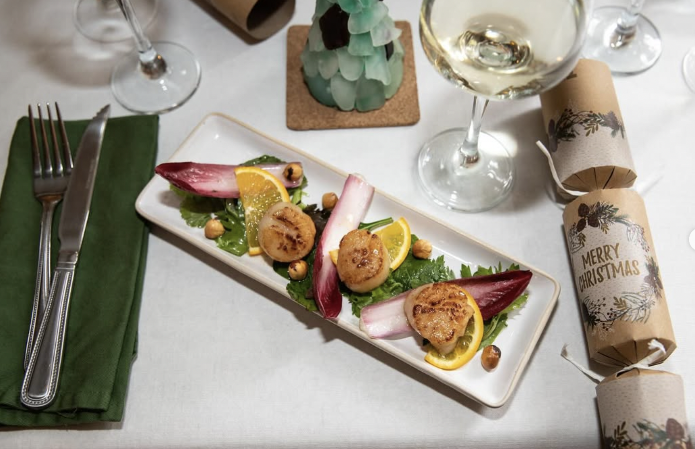 A holiday table setting with a rectangular plate of salad, two glasses of white wine, Christmas crackers, and a decorated Christmas tree ornament.