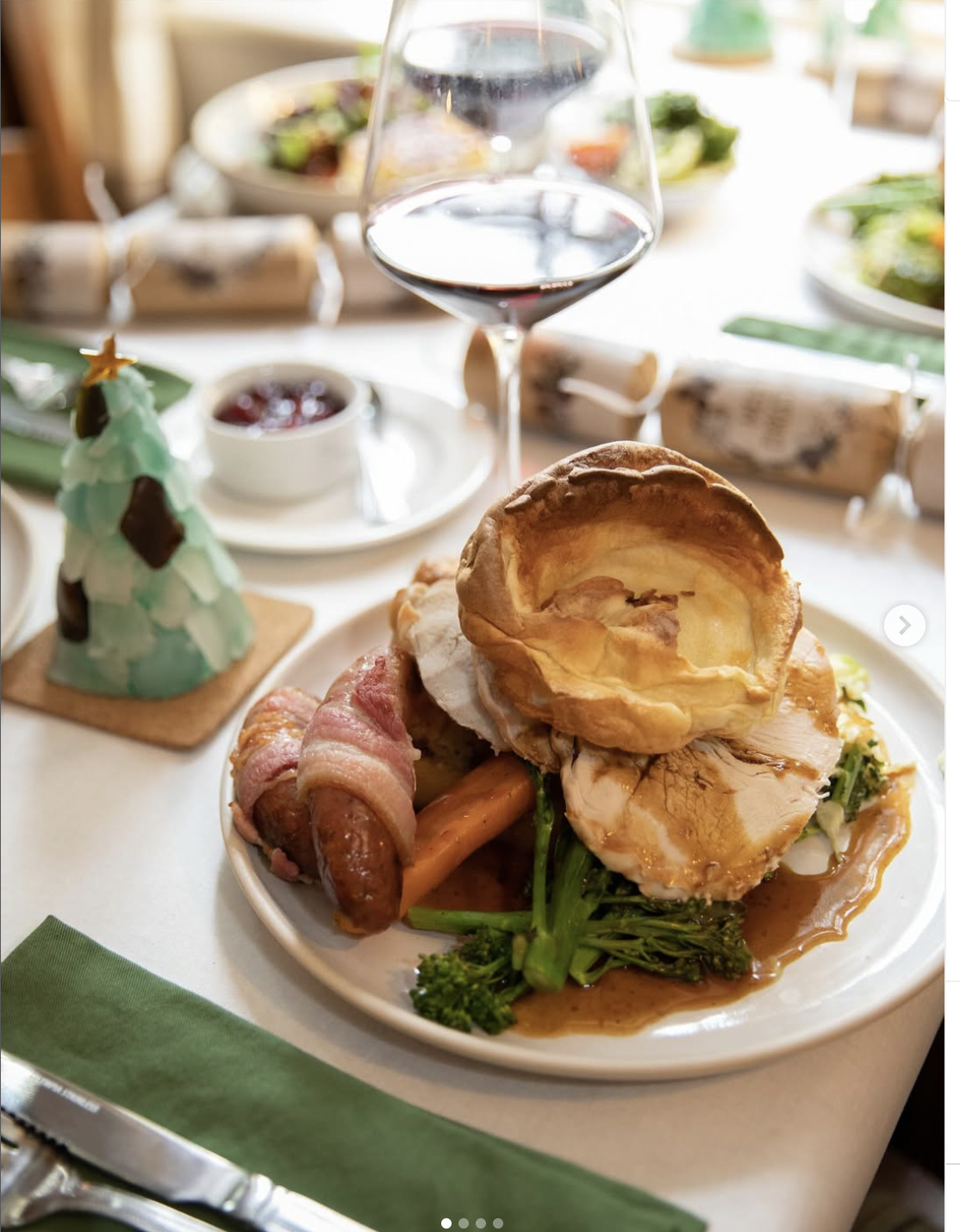 A plate with turkey, gravy, broccoli, bacon-wrapped sausage, and a Yorkshire pudding, with a glass of red wine on a decorated holiday table.