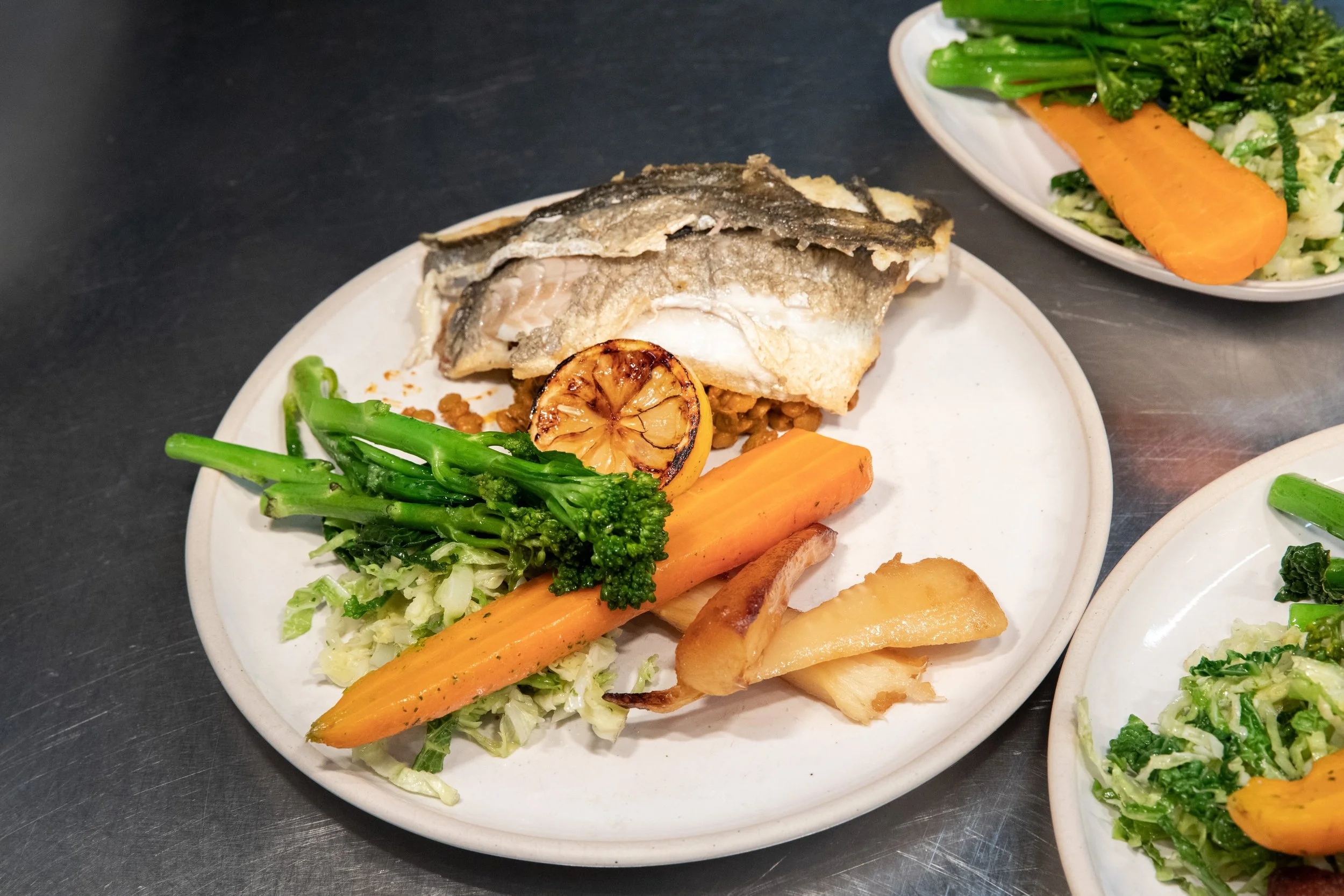Plate of cooked fish with vegetables including carrots, broccoli, and grilled lemon half. Another similar plate with vegetables visible in the background.