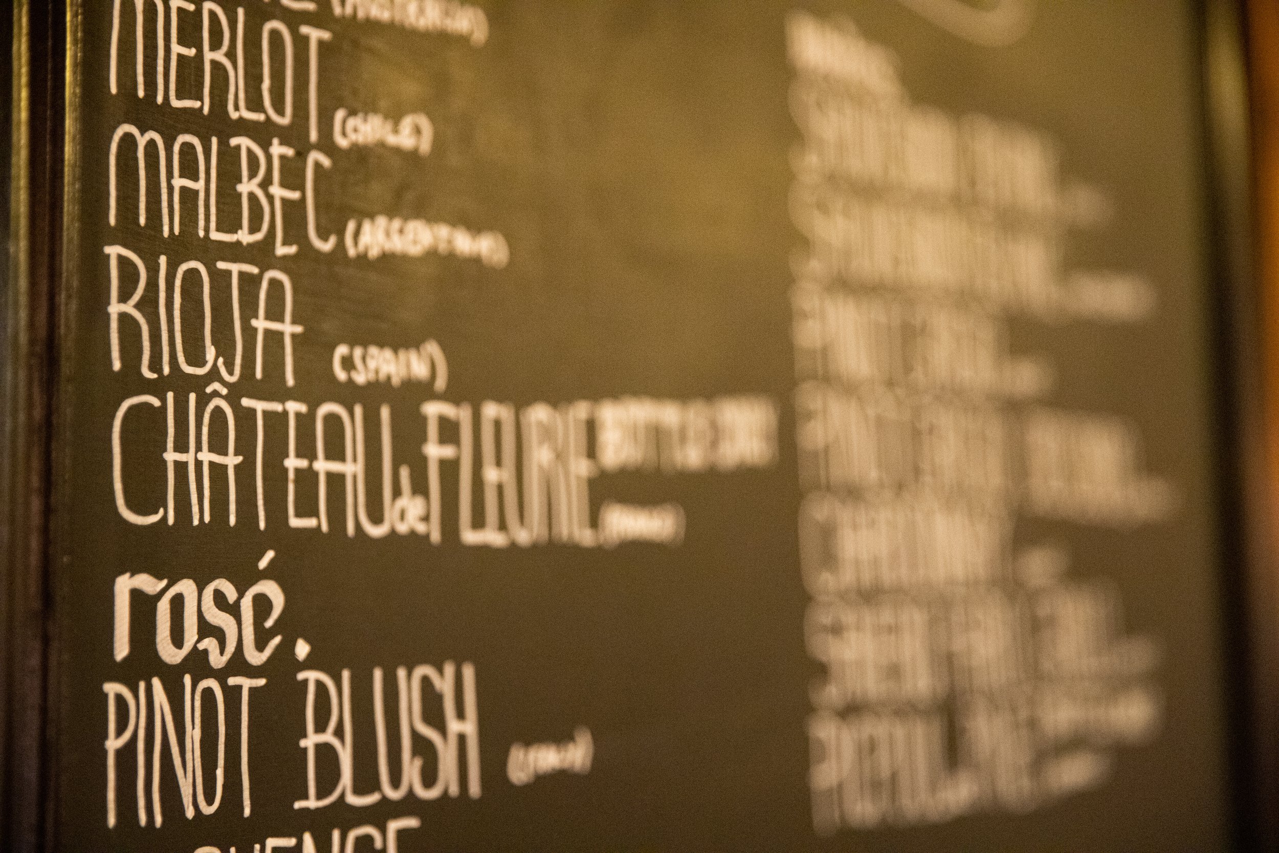 A menu board listing various wines such as Merlot, Malbec, Rioja, Châteauneuf-du-Pape, Rosé, Pinot Bouch, and Sauvignon, written in white chalk on a dark background.